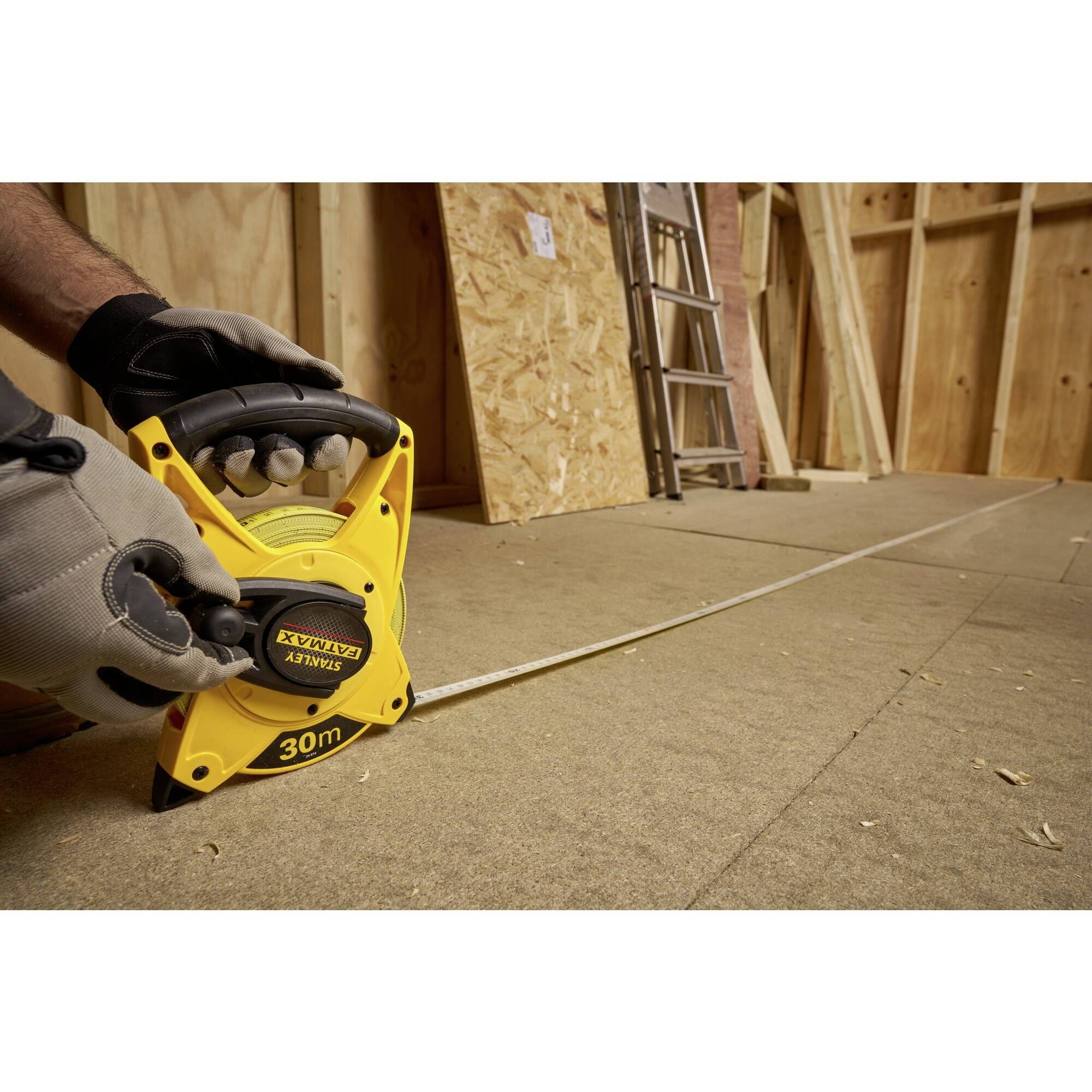 Person zieht ein gelbes Maßband in einer Baustelle aus, um den Abstand auf dem Boden zu messen. Eine Holzleiter ist an der Wand.