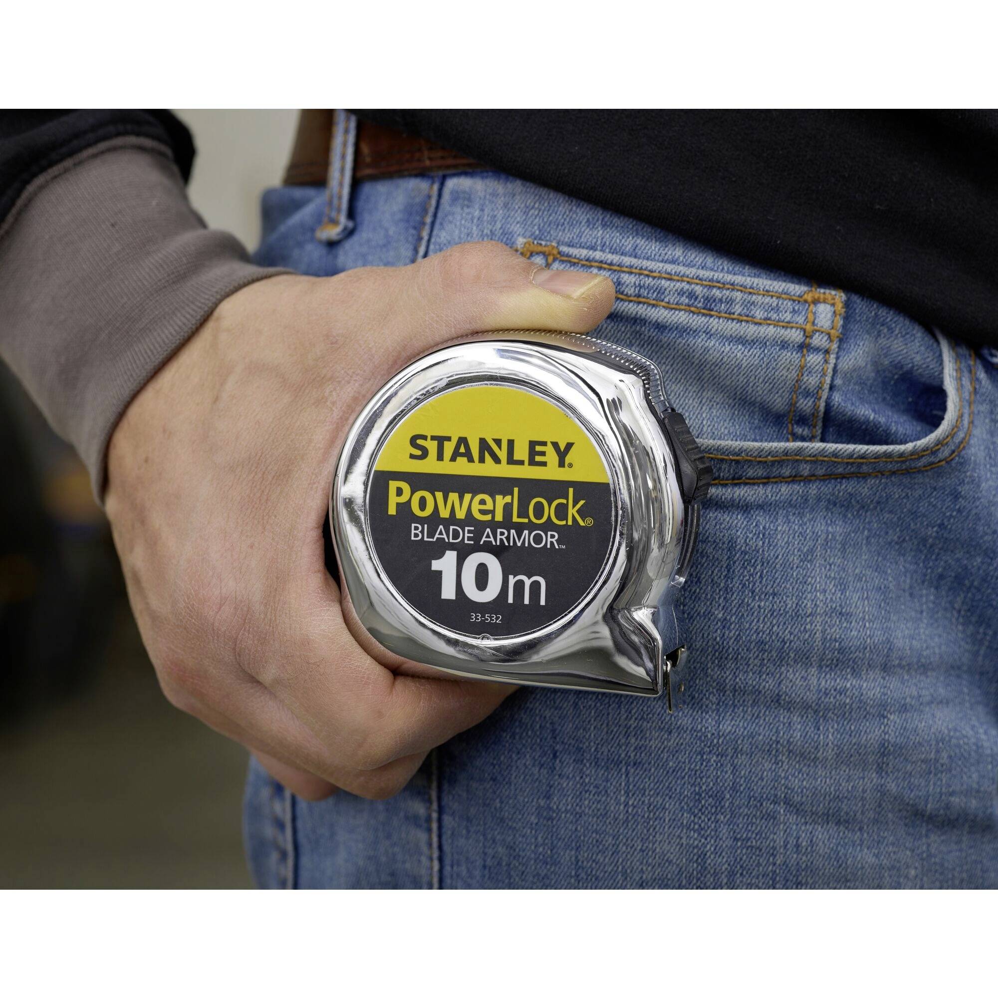 Eine Person hält ein Maßband mit der Aufschrift 'Stanley PowerLock Blade Armor 10m' in der rechten Hand. Das Maßband ist in der Nähe einer Hosentasche.