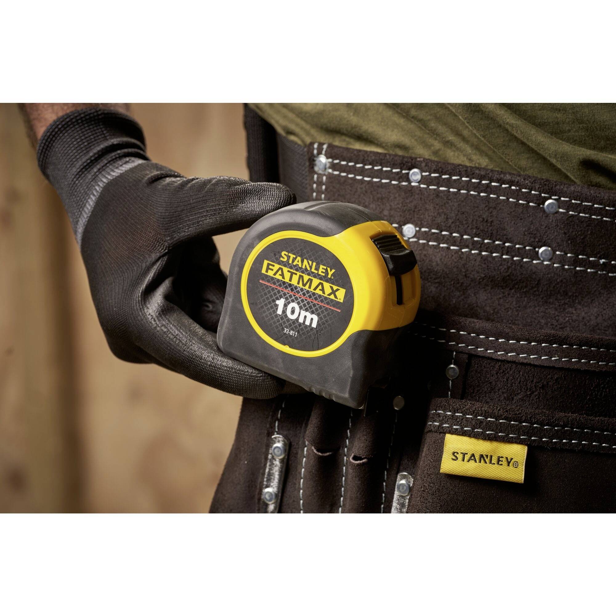 Eine bekleidete Hand hält ein 10m langes Stanley FatMax-Maßband. Im Hintergrund ist ein dunkelbrauner Werkzeuggürtel sichtbar.