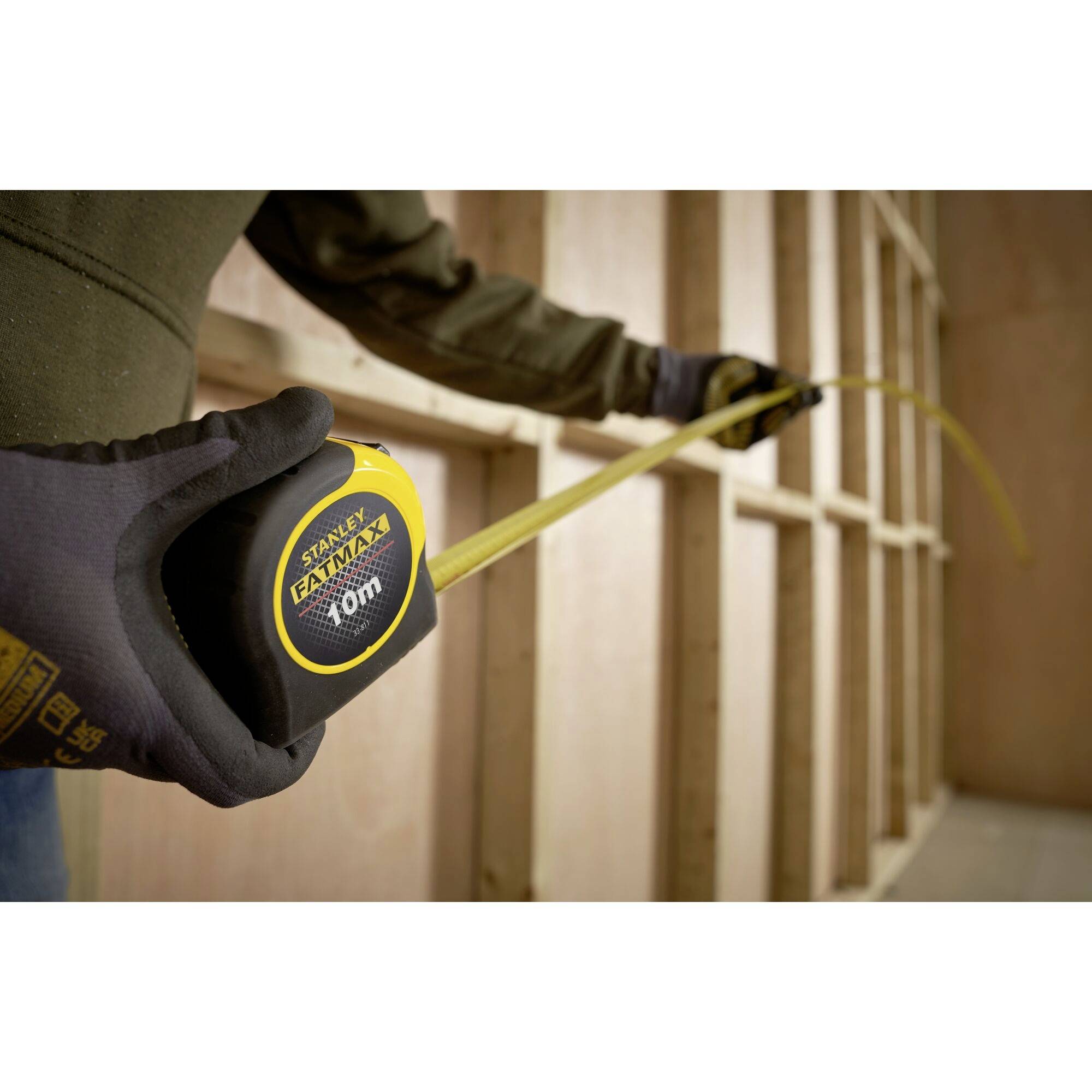 Eine Person mit Handschuhen misst mit einem Maßband Holzlatten. Das Maßband zeigt 10 Meter und ist in einer unvollendeten Wand.