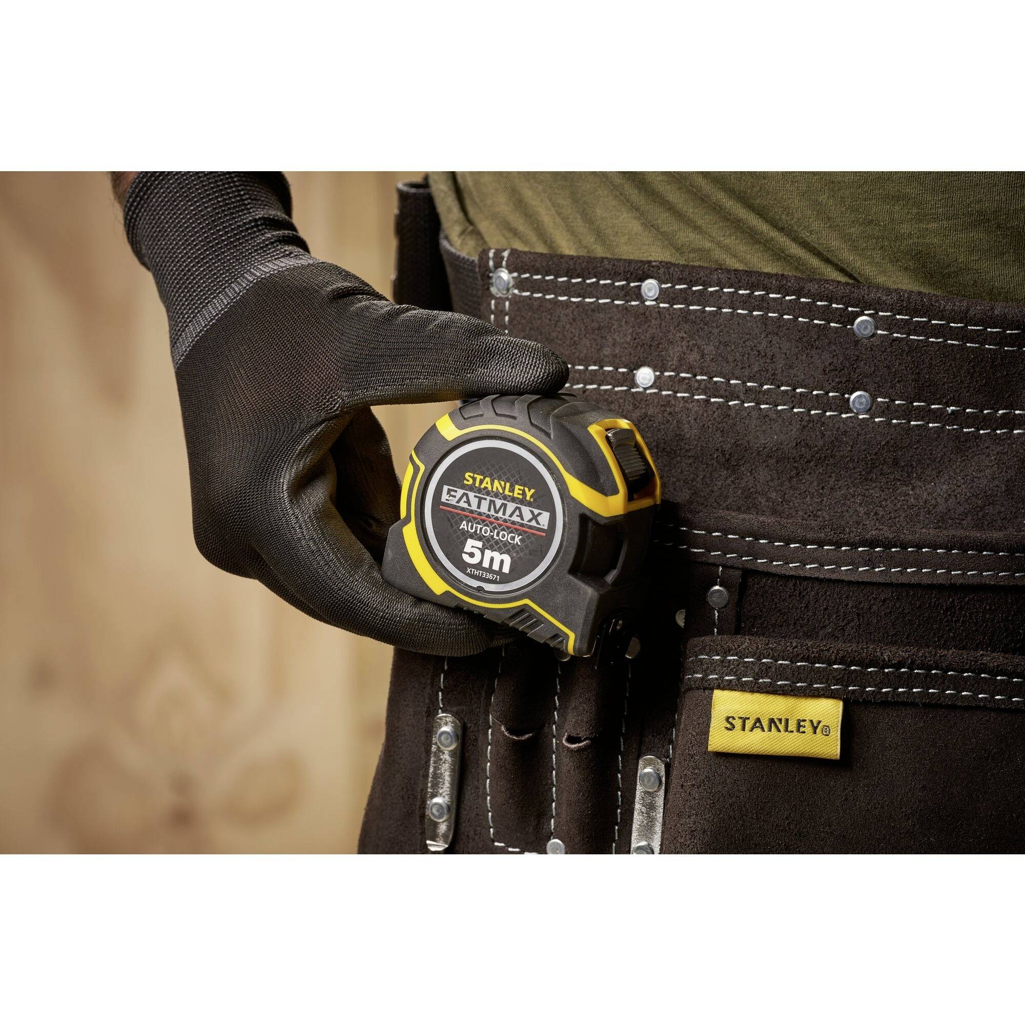 Eine Person trägt einen schwarzen Handschuh und hält ein gelbes Maßband der Marke Stanley FatMax, das in einem Arbeitsgürtel steckt.