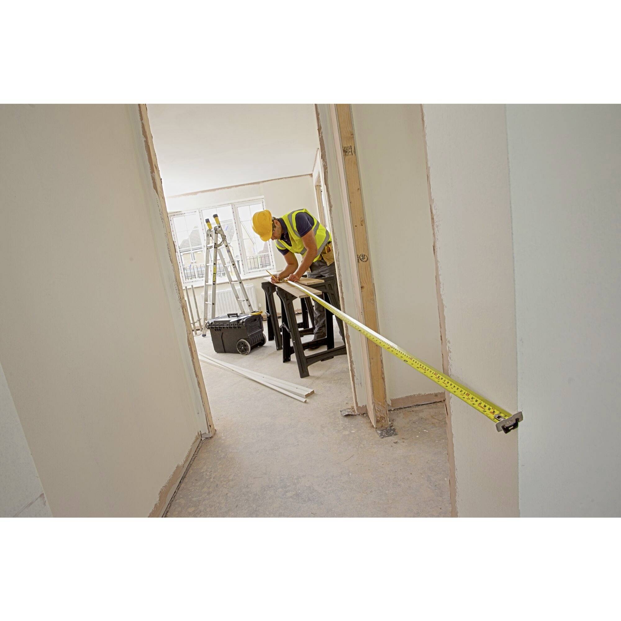 Zwei Bauarbeiter in Schutzhelmen messen mit einem langen Maßband in einem leeren Raum. Im Hintergrund ist Werkzeug sichtbar.