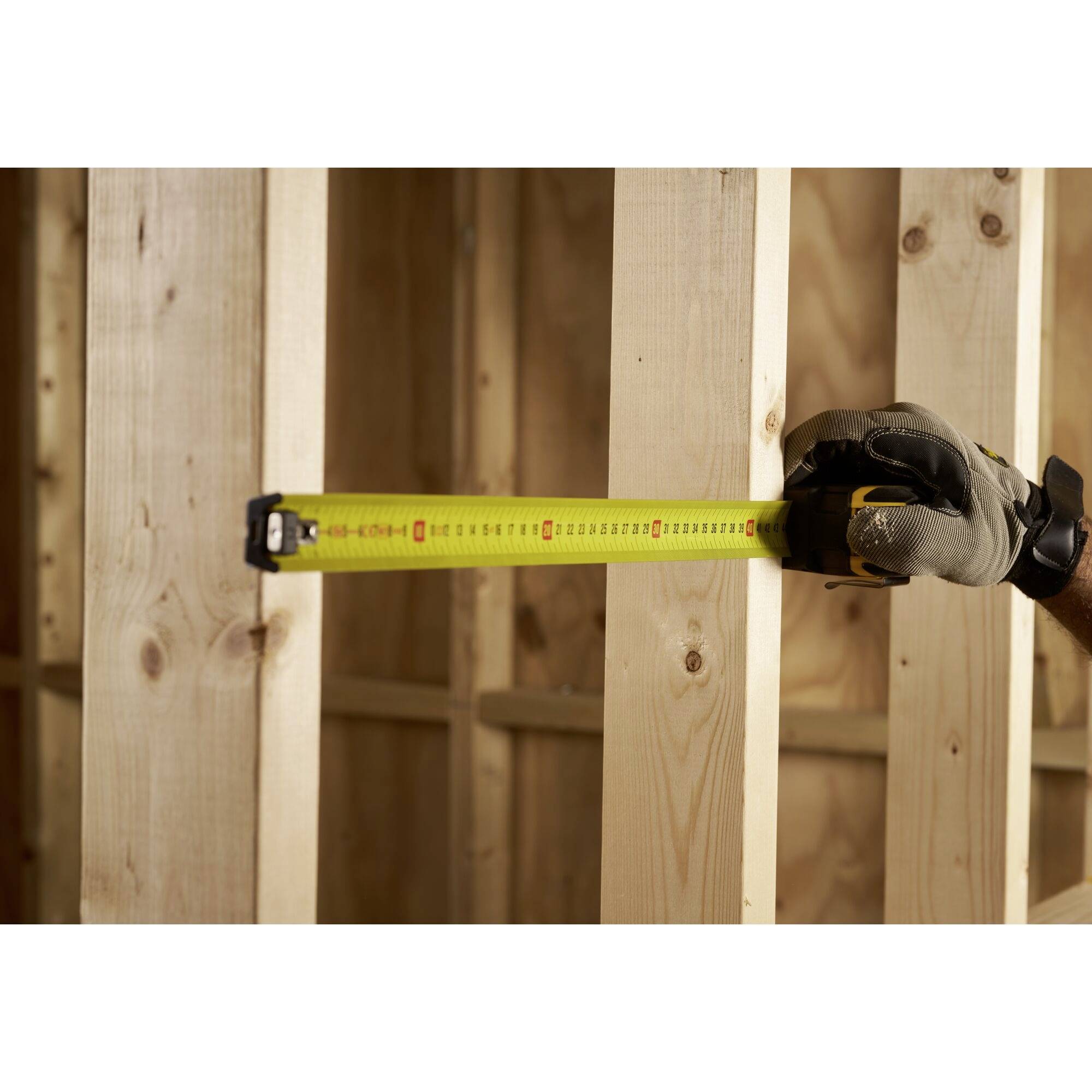 Ein Bauarbeiter hält ein ausgerolltes Maßband an einem Holzrahmen, misst die Breite zwischen zwei Holzbalken in einem Gebäude.