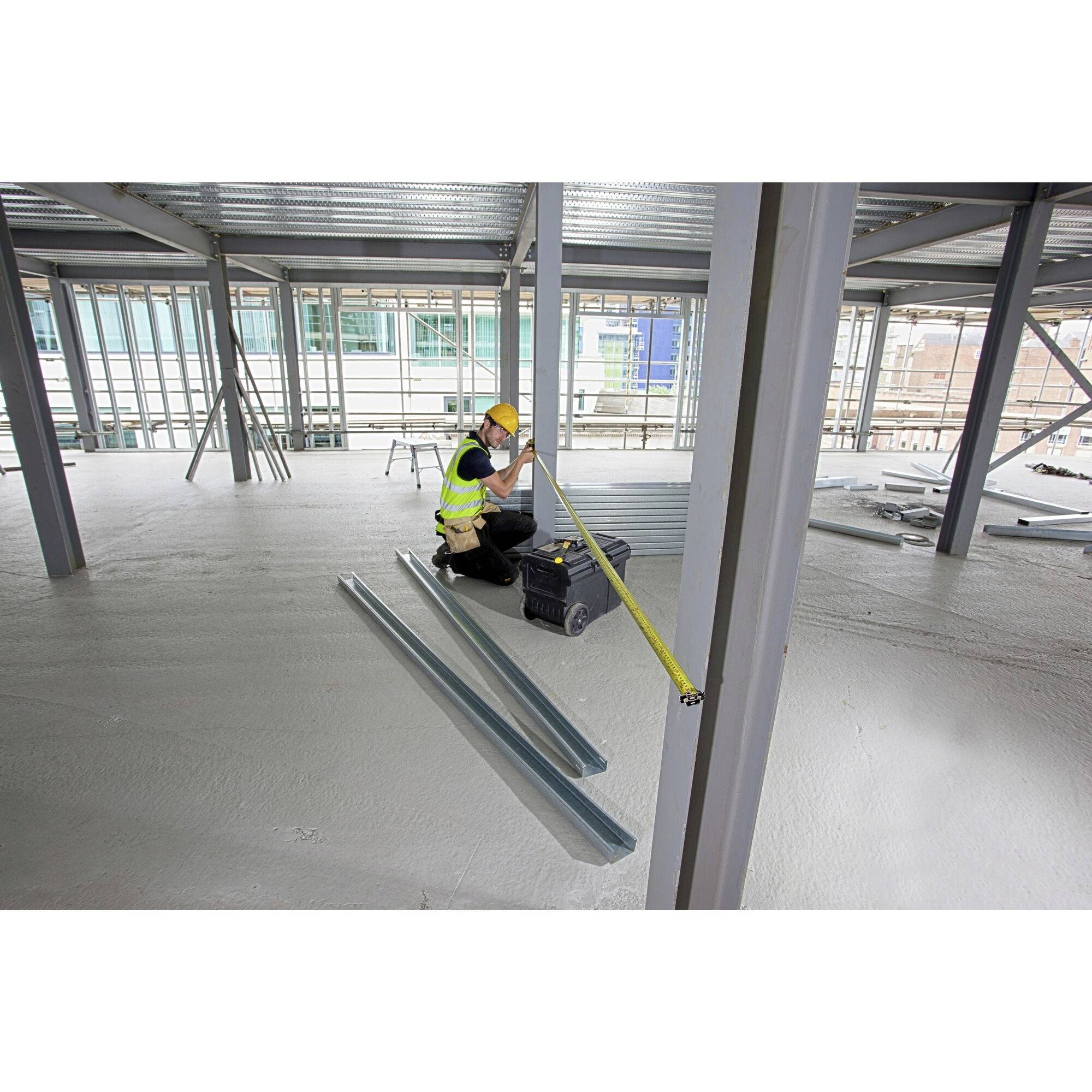 'Arbeiter misst Metallbalken auf einer Baustelle. Er trägt Schutzkleidung und Helm. Hintergrund zeigt Gebäudegerüst und Baustellenmaterialien.'