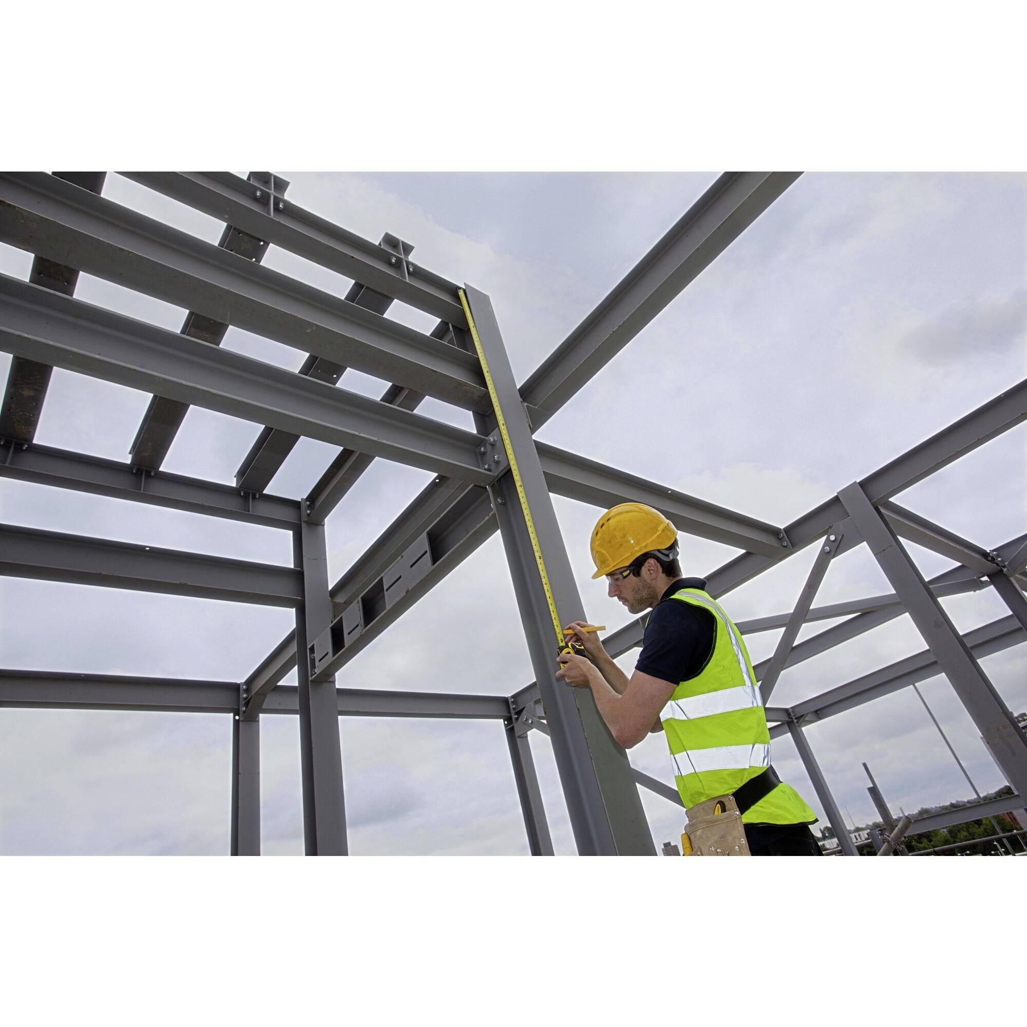 Ein Bauarbeiter mit gelbem Schutzhelm und Sicherheitsweste misst mit einem Maßband die Höhe eines Stahlträgers auf einer Baustelle.