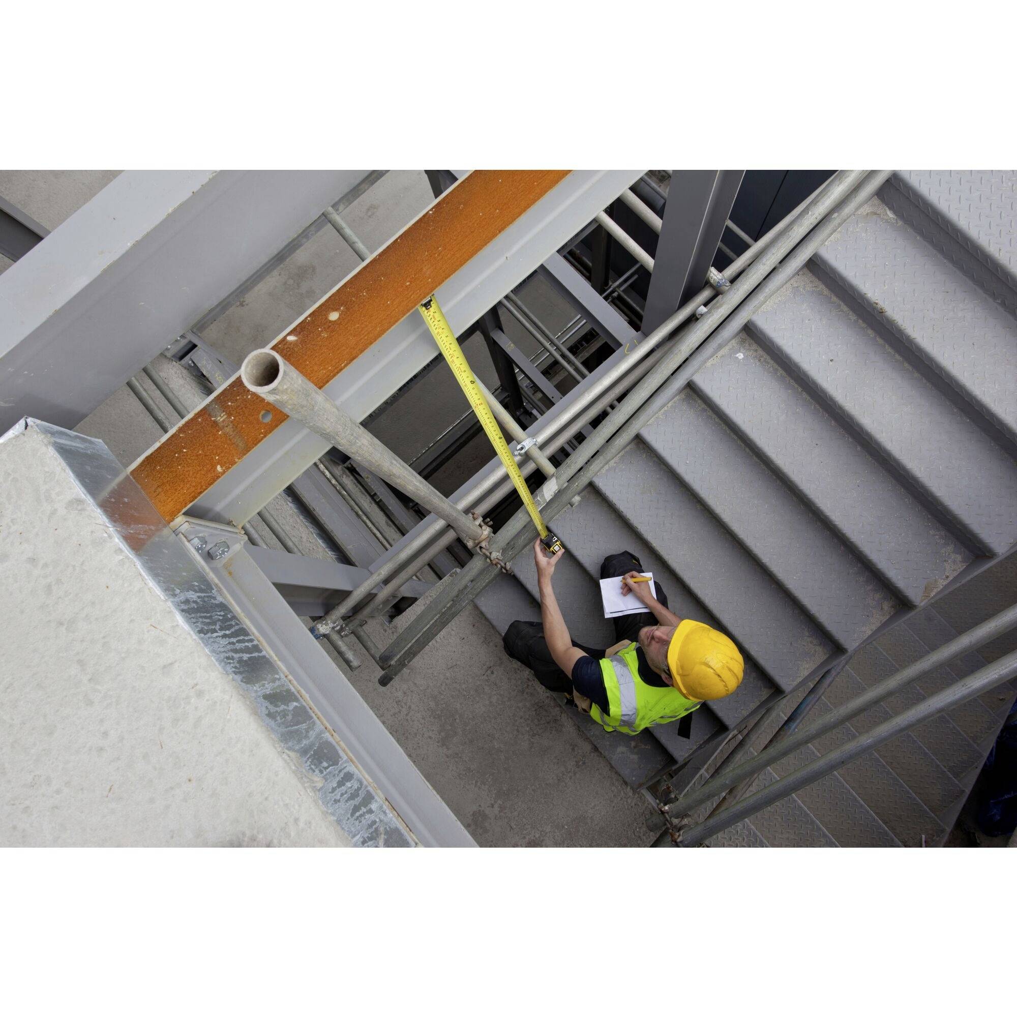 Ein Arbeiter mit gelbem Helm und Warnweste misst mit einem Maßband eine Metalltreppe und notiert Ergebnisse in ein Notizbuch.