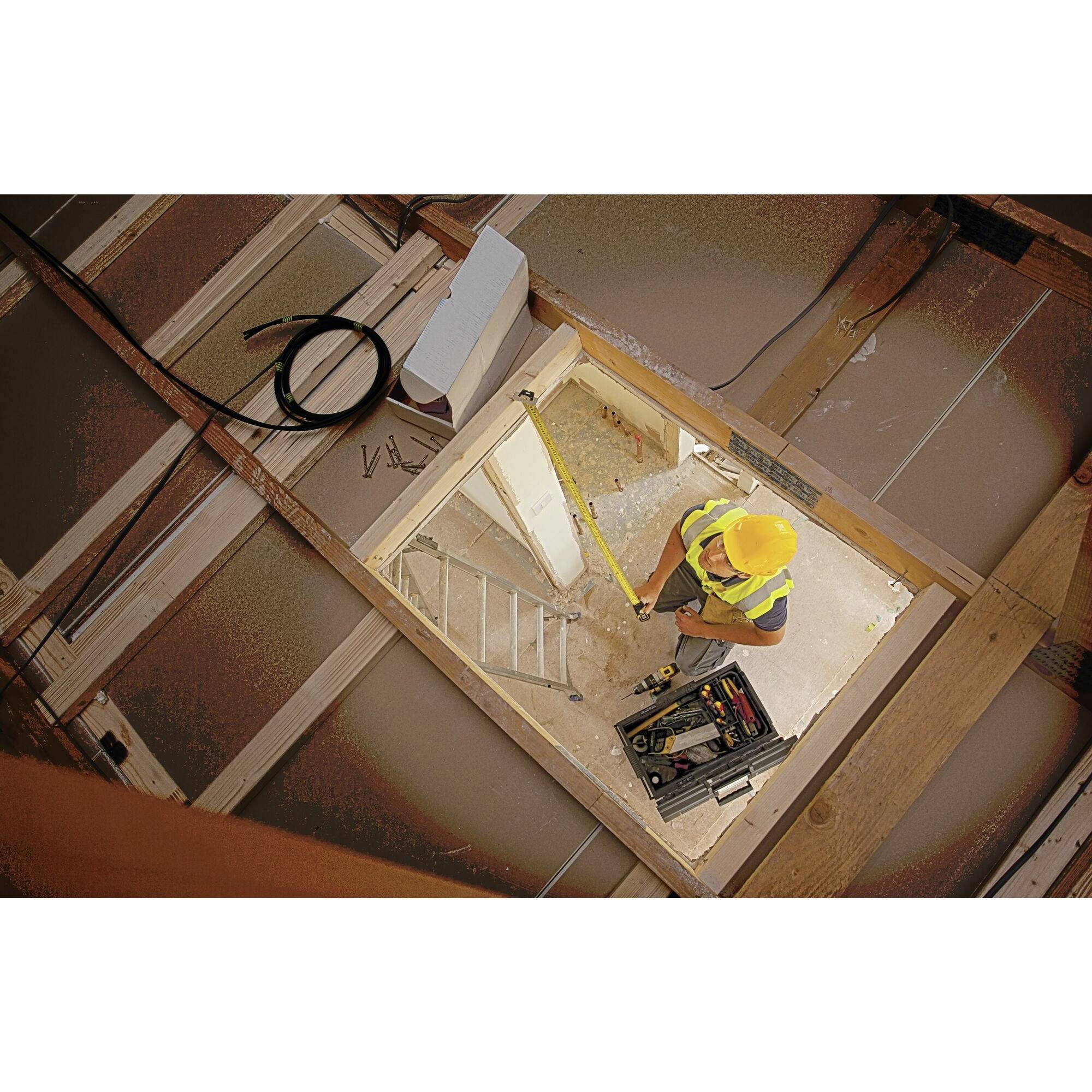 Ein Bauarbeiter mit Schutzhelm arbeitet in einem Dachboden. Vor ihm liegen Werkzeuge und Kabel. Der Raum ist noch unvollständig ausgebaut.