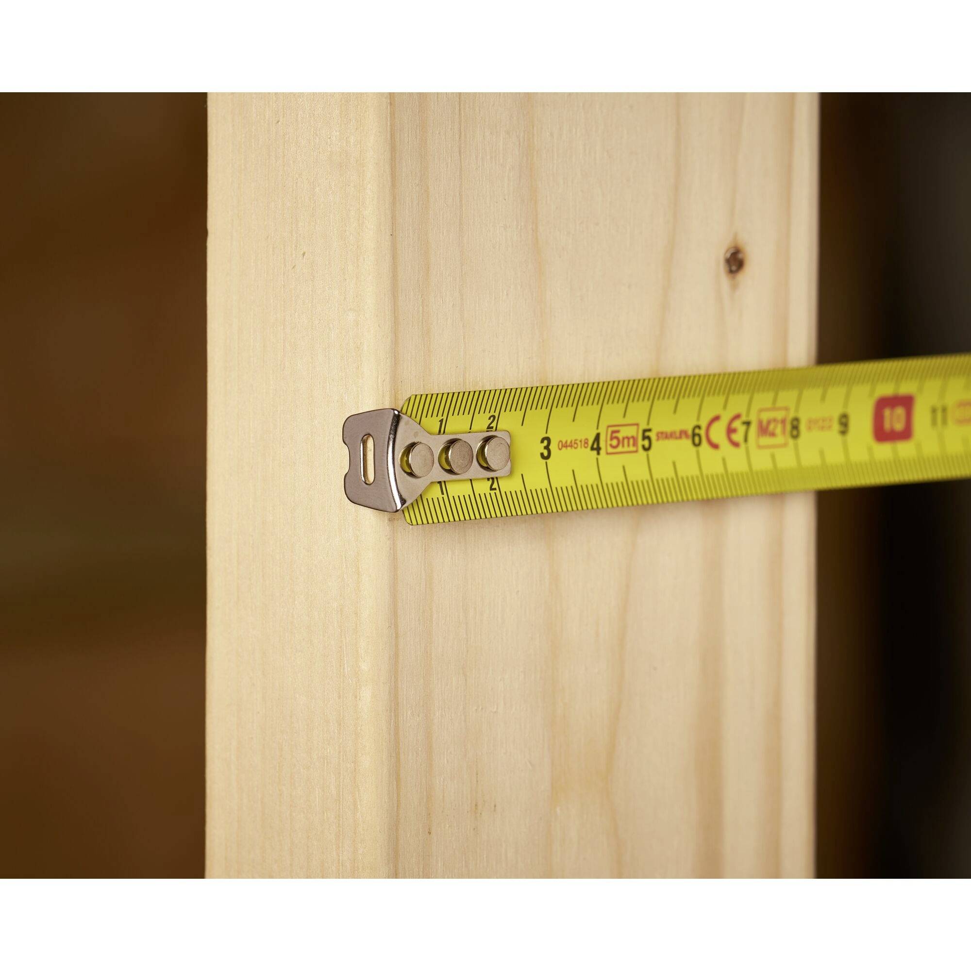 Ein Maßband misst die Breite eines Holzbalkens. Das Band zeigt 10 cm an. Der Hintergrund ist unscharf.