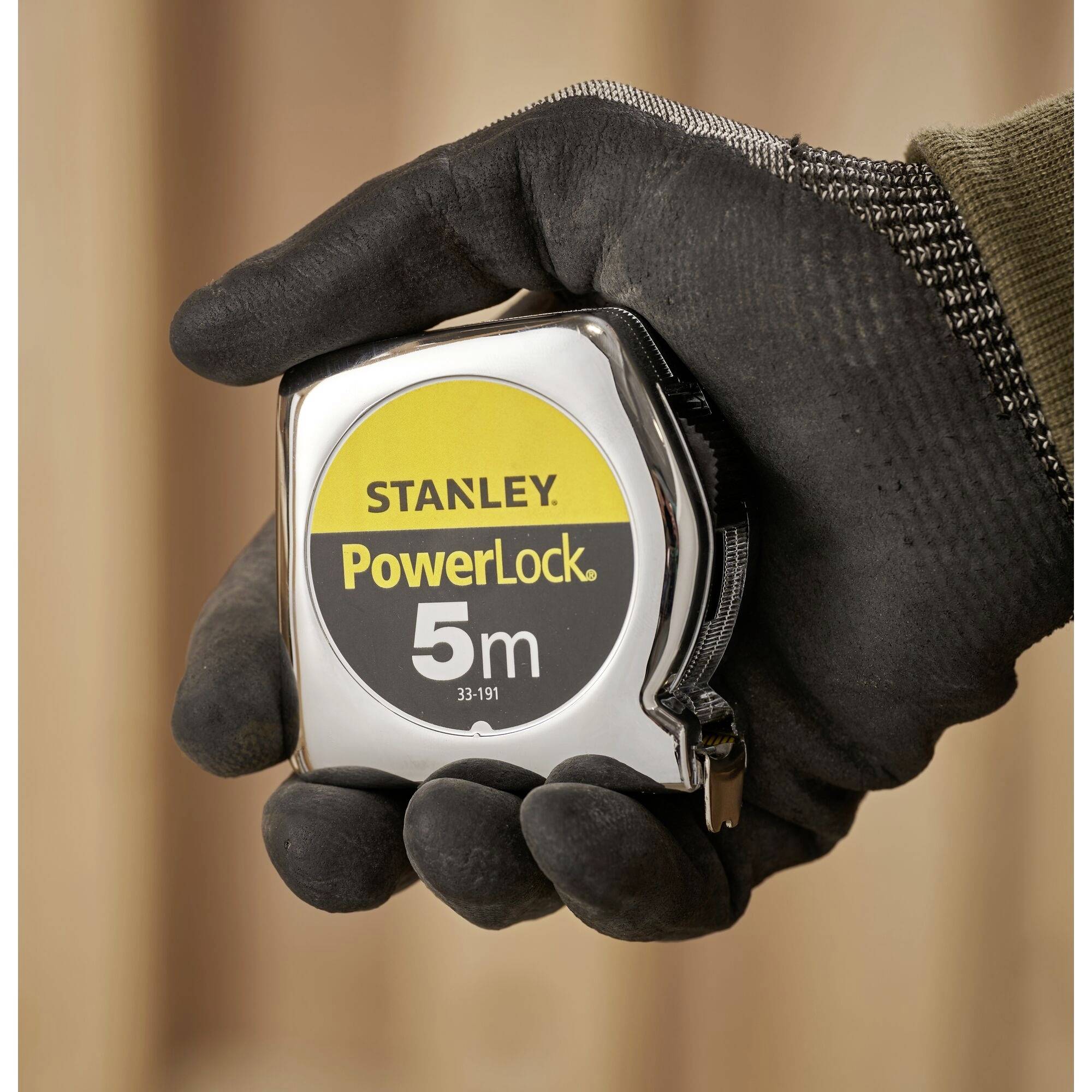 Eine Hand in einem Handschuh hält ein Stanley PowerLock 5m Maßband. Der Hintergrund ist unscharf.