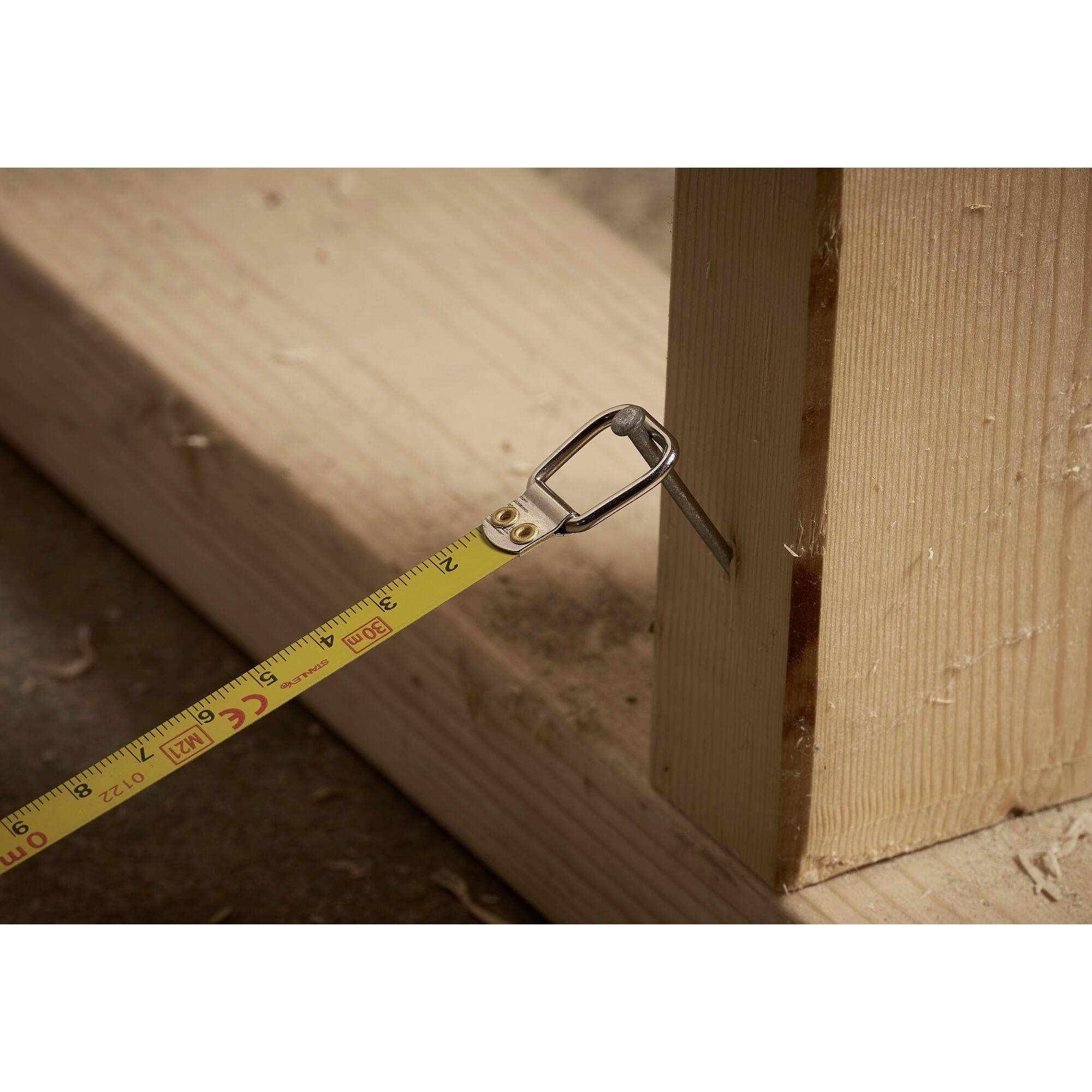 Ein gelbes Maßband misst die Breite eines Holzbalkens. Der Hintergrund zeigt eine Werkstattumgebung mit Holzspänen.