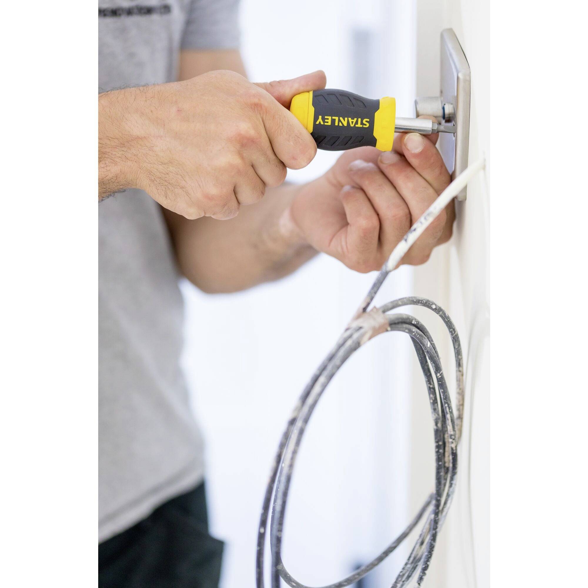 Eine Person repariert eine Steckdose an einer weißen Wand mit einem Schraubenzieher. Kabel hängen unter der Steckdose hervor.