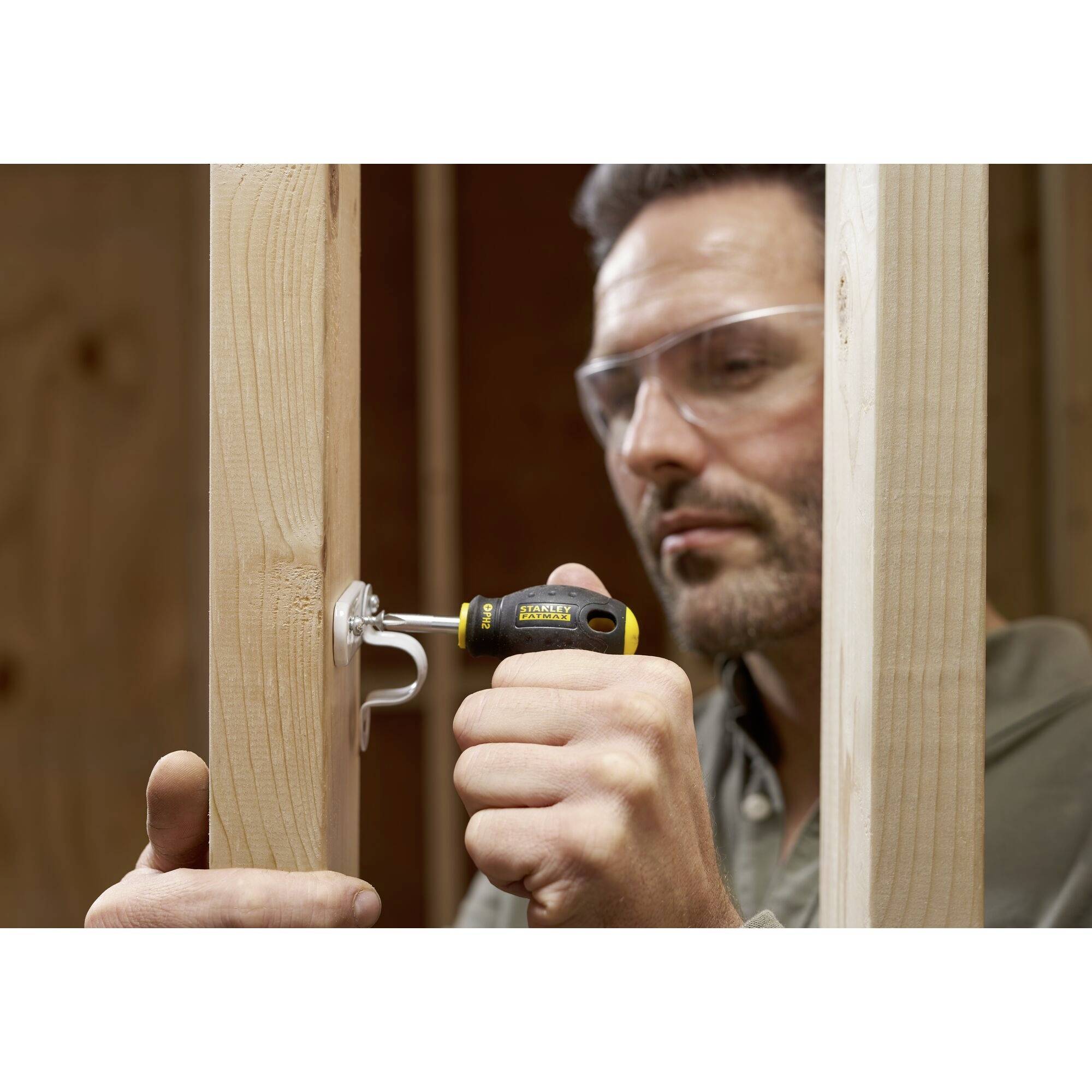 Ein Mann mit Schutzbrille benutzt einen gelben Schraubenzieher, um eine Schraube in einen Holzrahmen zu drehen.