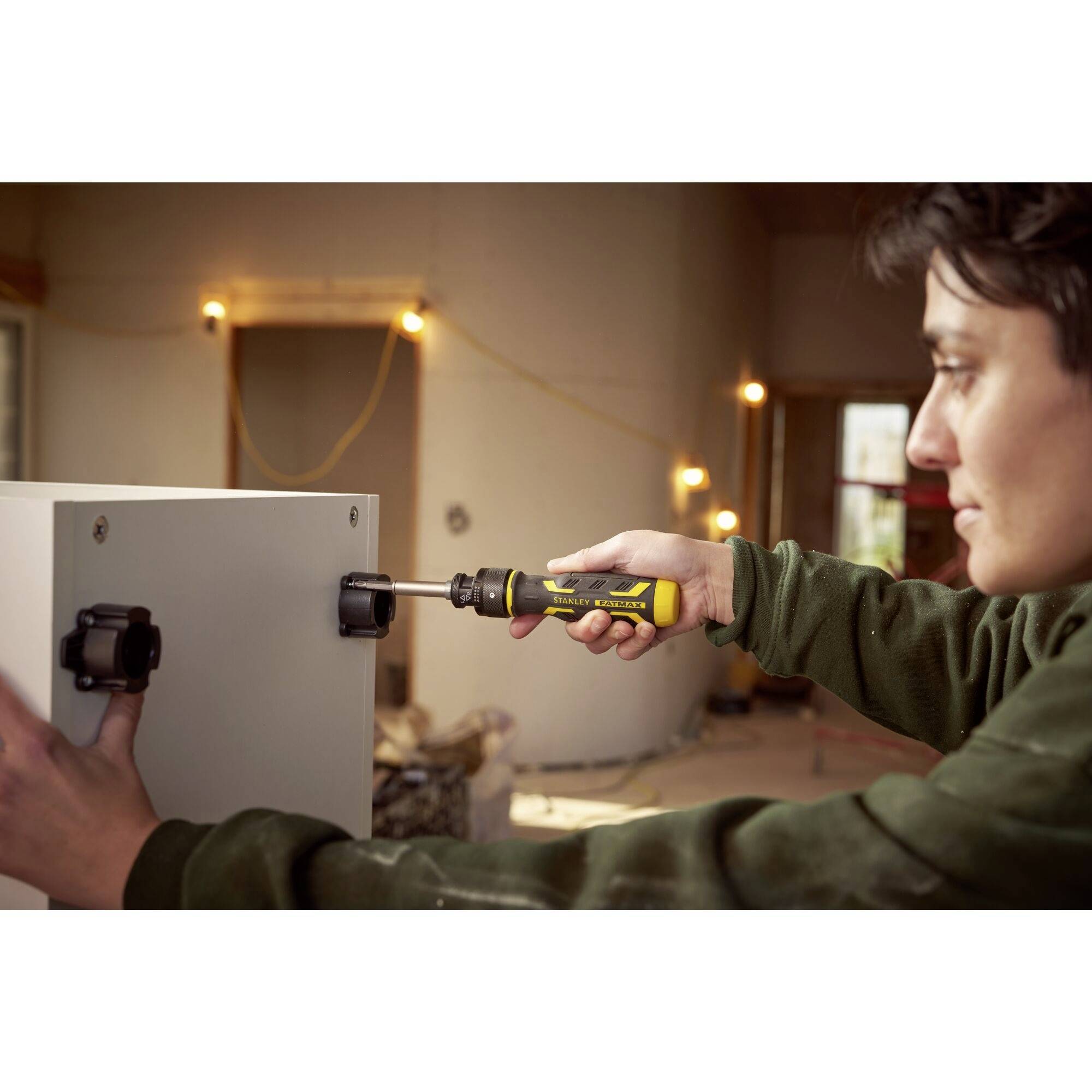 Person montiert ein weißes Möbelstück mit einem Schraubenzieher in einem Raum, der sich im Umbau befindet. Im Hintergrund sind Baulichter.