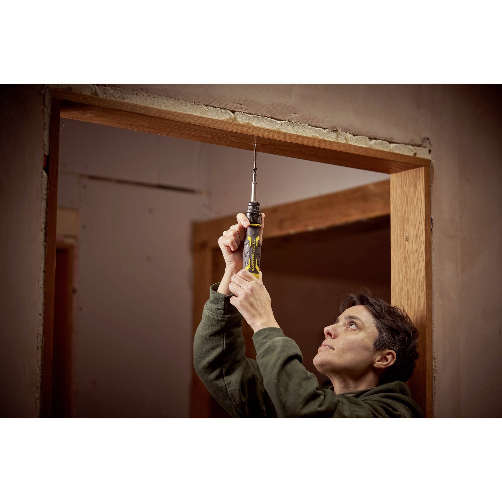 Person in einem Raum verwendet einen gelben Schraubendreher, um in den oberen Türrahmen einer unvollendeten Holzwand zu schrauben.