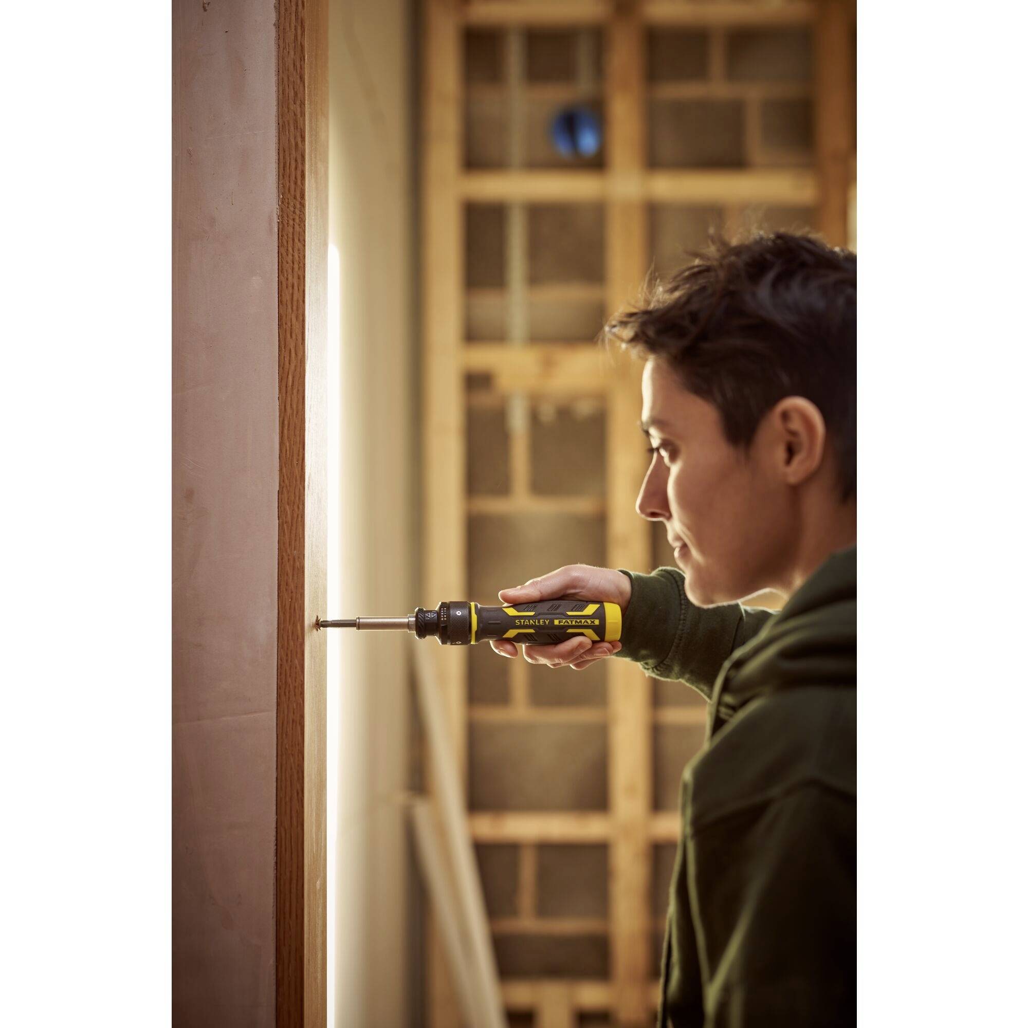 Eine Person verwendet einen Schraubendreher, um eine Schraube in einen Holzbalken an einer Wand zu befestigen. Im Hintergrund ist eine Baustelle sichtbar.