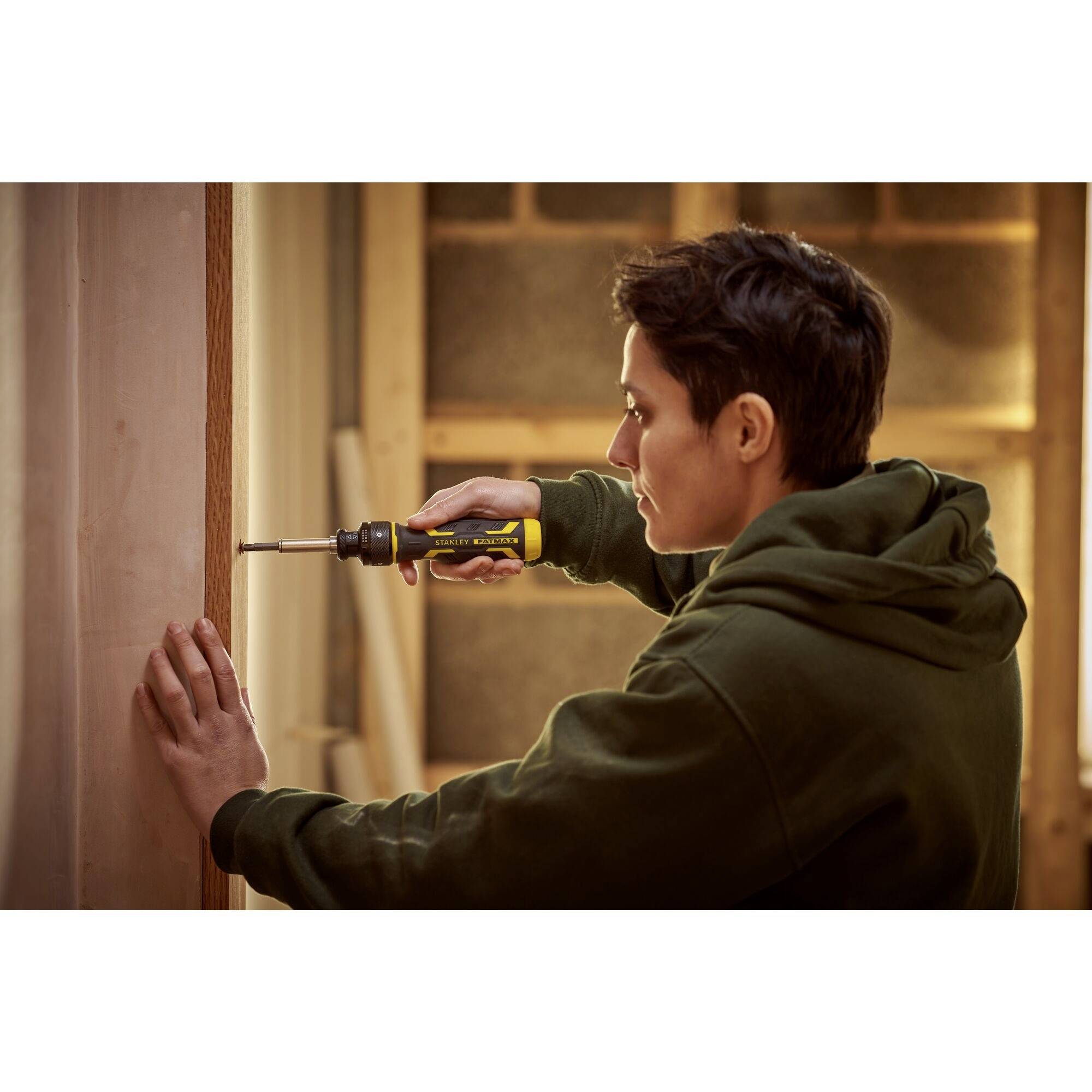 Eine Person verwendet einen Akkuschrauber, um eine Schraube in eine Holzwand zu drehen. Holzplatten und Werkzeuge im Hintergrund.