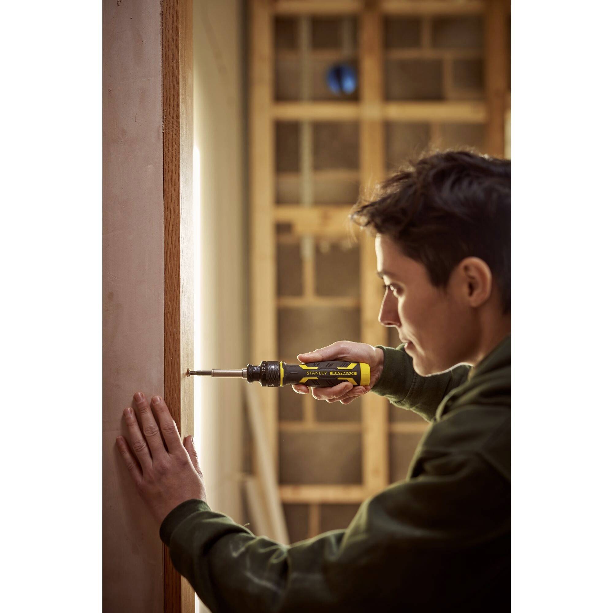 Eine Person verwendet einen Schraubendreher, um ein Stück Holz in einem teilweise fertiggestellten Raum zu befestigen. Im Hintergrund sind Holzbalken sichtbar.