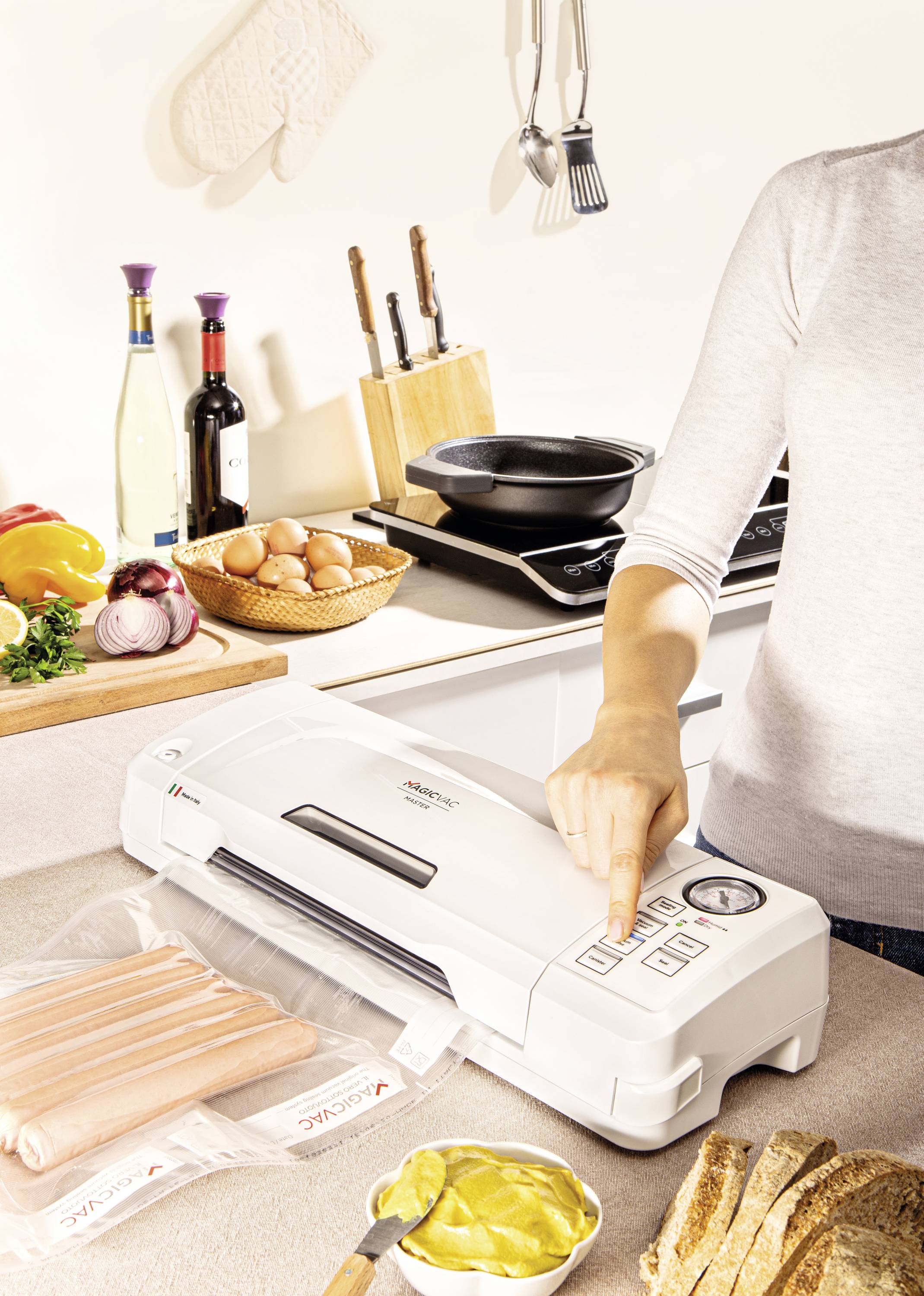 Person bedient ein Vakuumiergerät in einer Küche. Es gibt Lebensmittel wie Brot, Wurst und Eier neben dem Gerät. Küchenutensilien im Hintergrund.