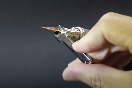 Eine Hand hält eine Tätowiermaschine mit sichtbarer Nadel, auf schwarzem Hintergrund.