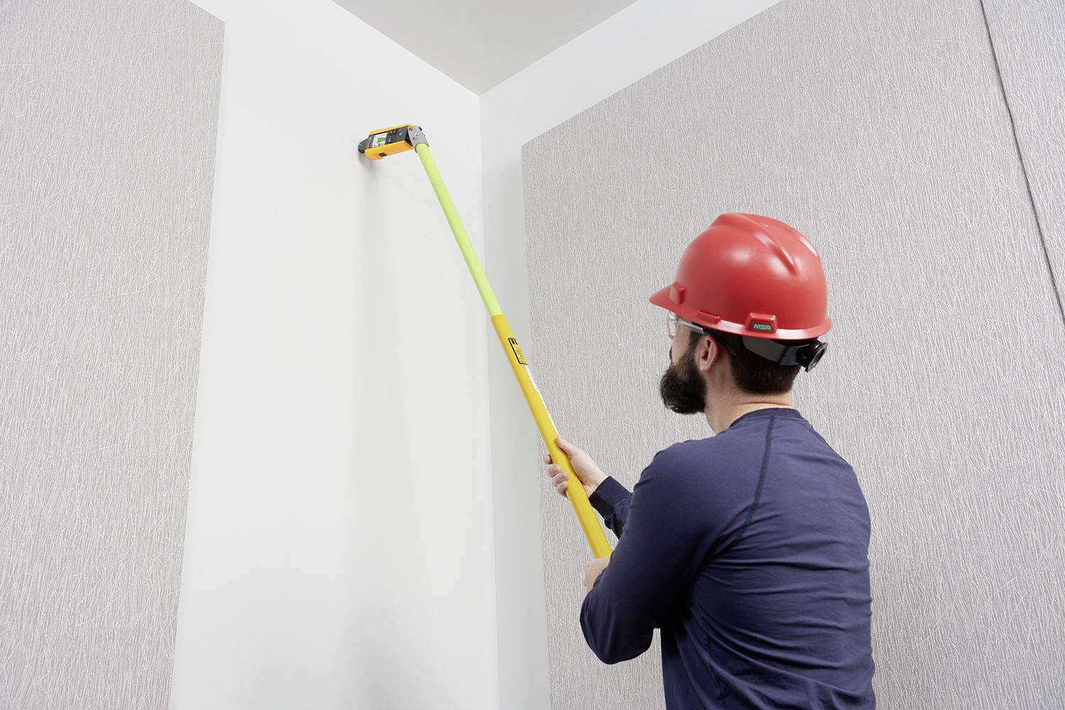 Ein Handwerker trägt einen roten Schutzhelm und benutzt eine gelbe Teleskopstange mit Werkzeug, um in einer Raumecke zu arbeiten.