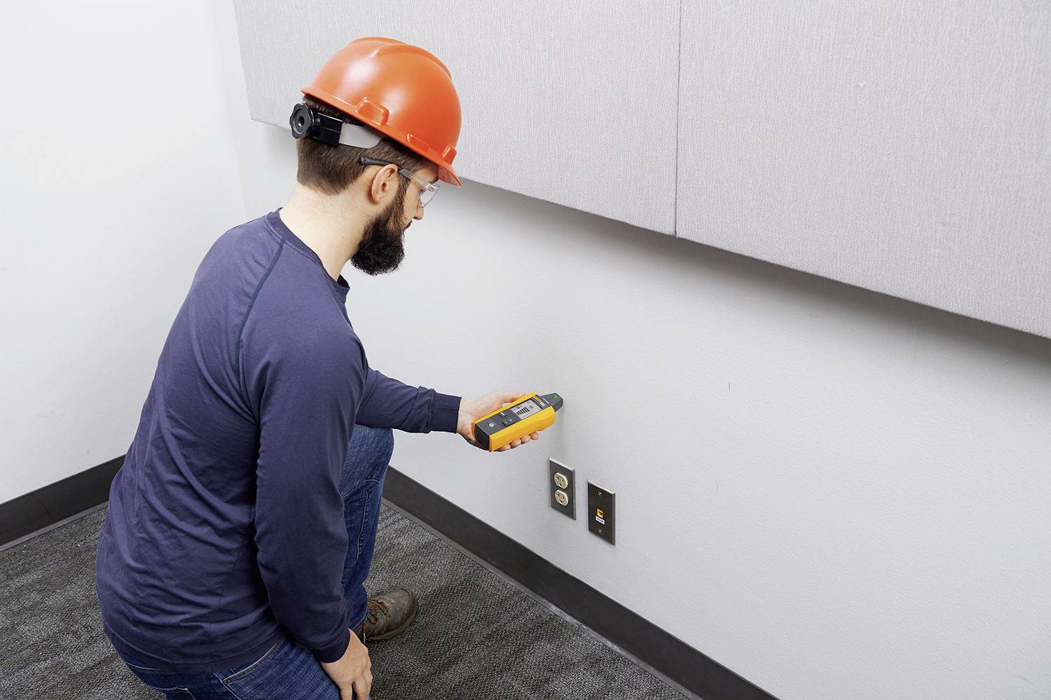 Ein Mann mit Schutzhelm überprüft eine Steckdose an einer Wand mit einem Prüfgerät.