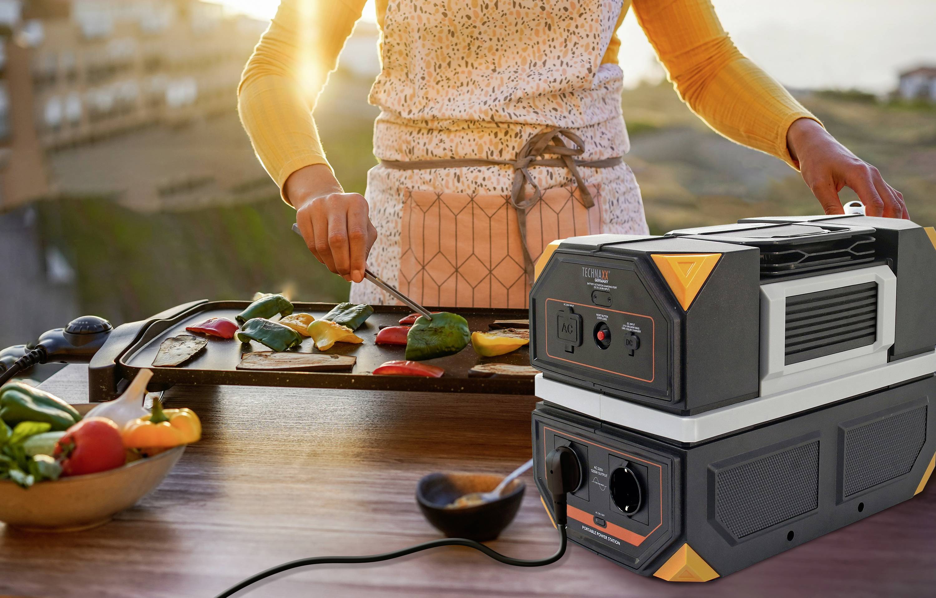 Eine Person grillt Gemüse auf einer Terrasse bei Sonnenuntergang. Im Vordergrund steht eine tragbare Stromstation, die angeschlossen ist.