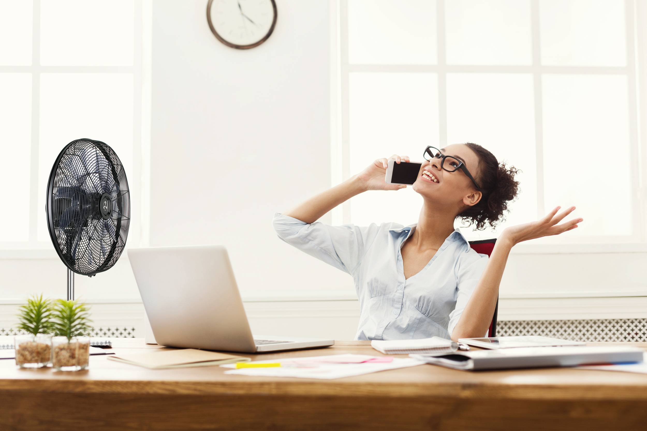 Eine Frau sitzt an einem Schreibtisch mit Laptop und spricht fröhlich am Smartphone. Neben ihr steht ein Ventilator.