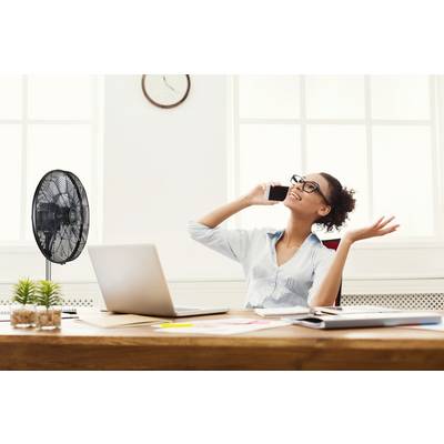 Eine Frau sitzt an einem Schreibtisch mit Laptop und spricht fröhlich am Smartphone. Neben ihr steht ein Ventilator.