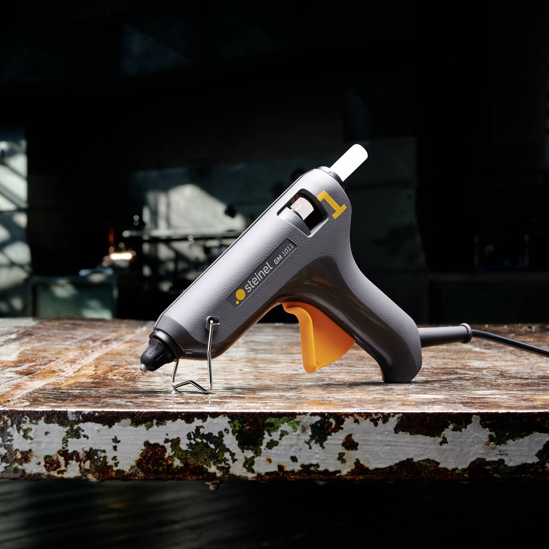 Eine Klebepistole liegt auf einem abgenutzten Holztisch. Im Hintergrund ist ein dunkler Raum sichtbar.