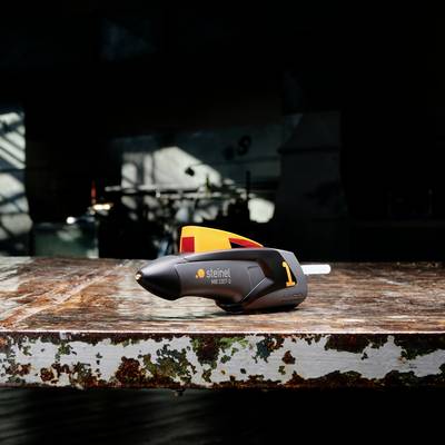 Eine Heißklebepistole der Marke Steinel liegt auf einem abgenutzten Tisch in einer Werkstatt. Im Hintergrund unscharf: Werkzeuge.