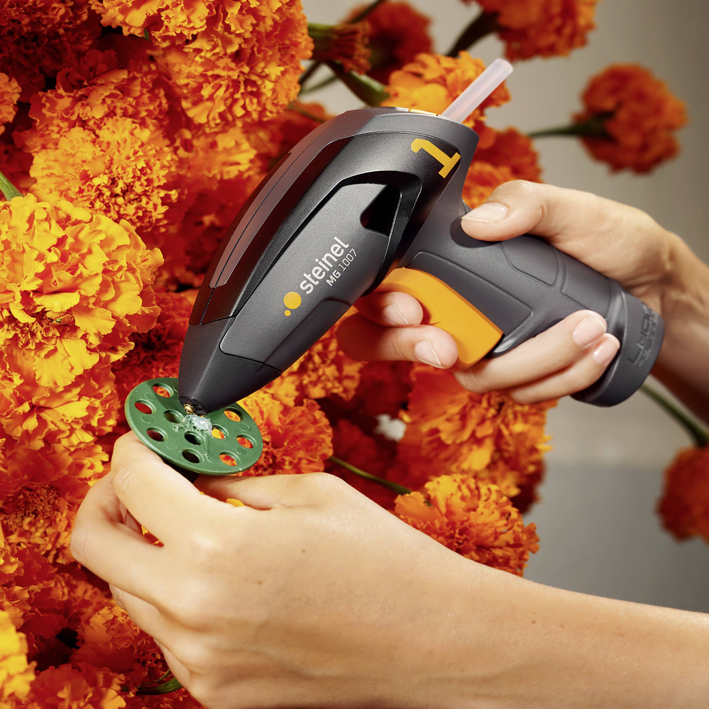 Eine Hand hält eine Heißklebepistole und klebt einen grünen Dekorationsring auf orangefarbene Blumen.