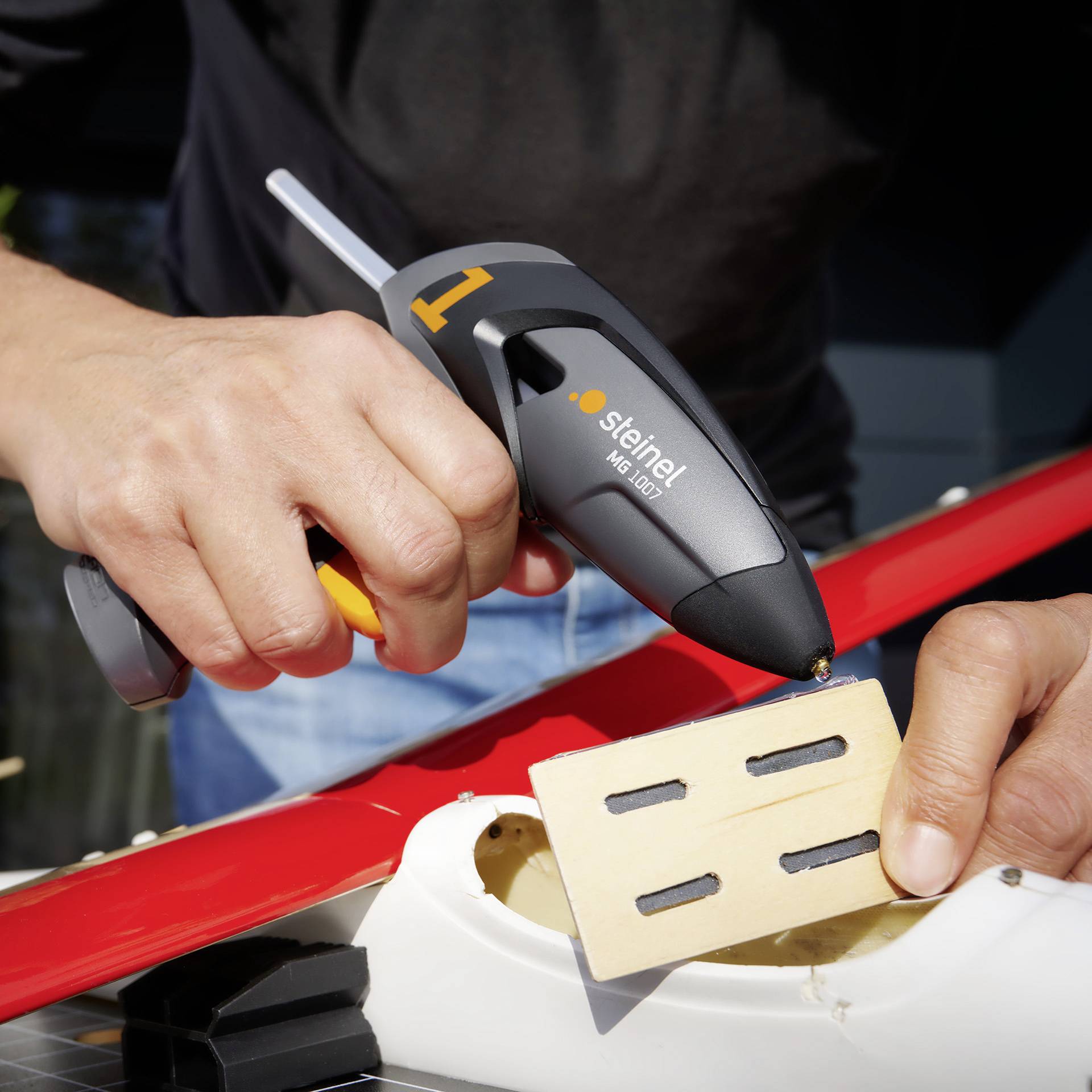 'Person verwendet eine Heißklebepistole, um ein Holzstück auf einem Modellflugzeug zu befestigen. Markenname ist sichtbar.'