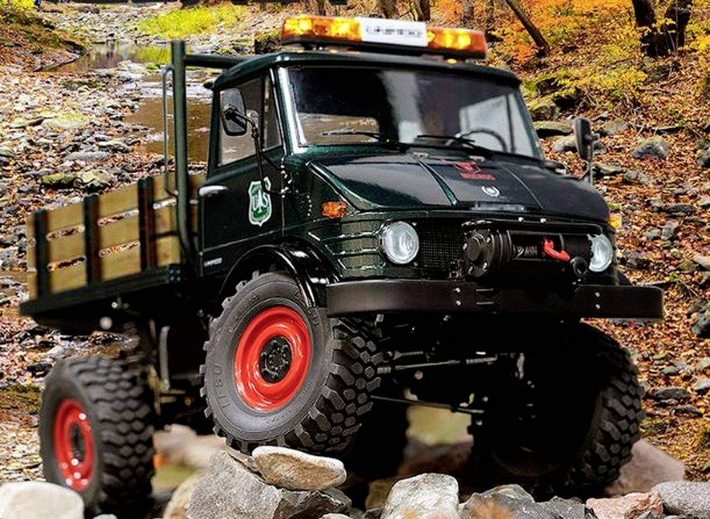Ein geländegängiger Lastwagen mit orangefarbenen Warnlichtern fährt über eine steinige, herbstliche Waldlandschaft.