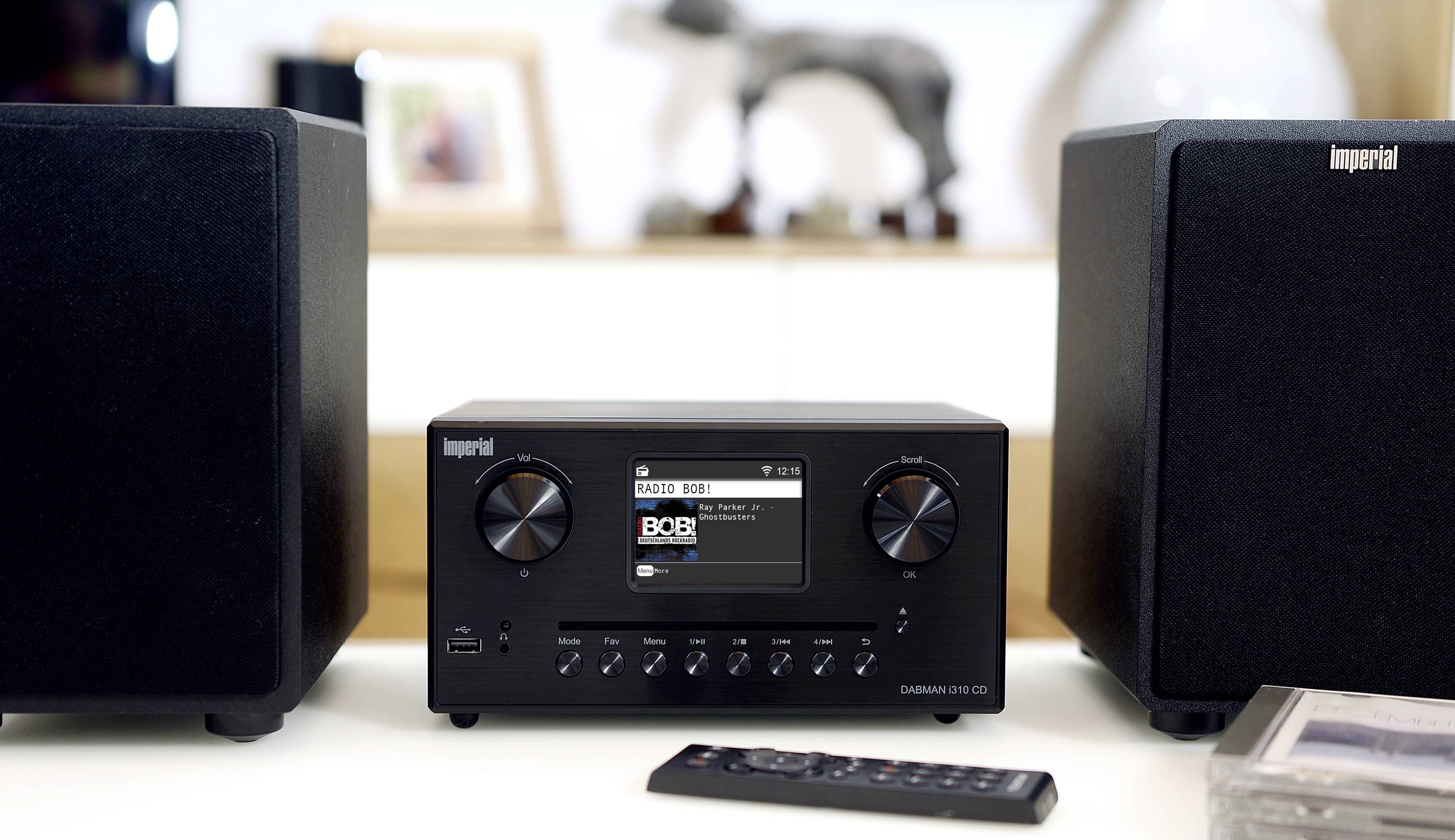 Schwarzes Audiosystem mit Display in der Mitte, umgeben von zwei Lautsprechern. Fernbedienung liegt auf dem Tisch davor.