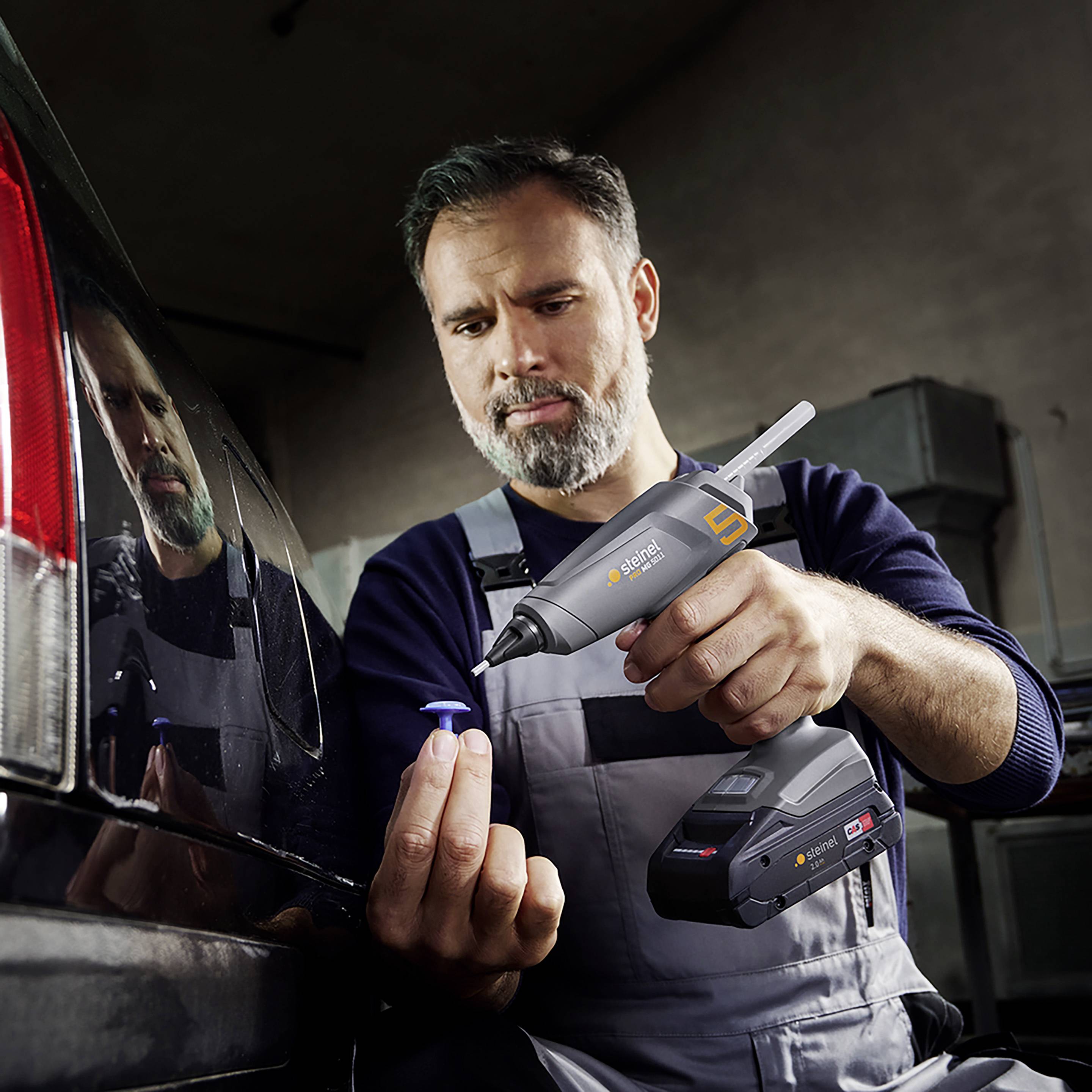 Ein Mechaniker verwendet ein tragbares Klebepistolen-Werkzeug, um ein Kunststoffteil an einem Auto zu befestigen. Er wirkt konzentriert.