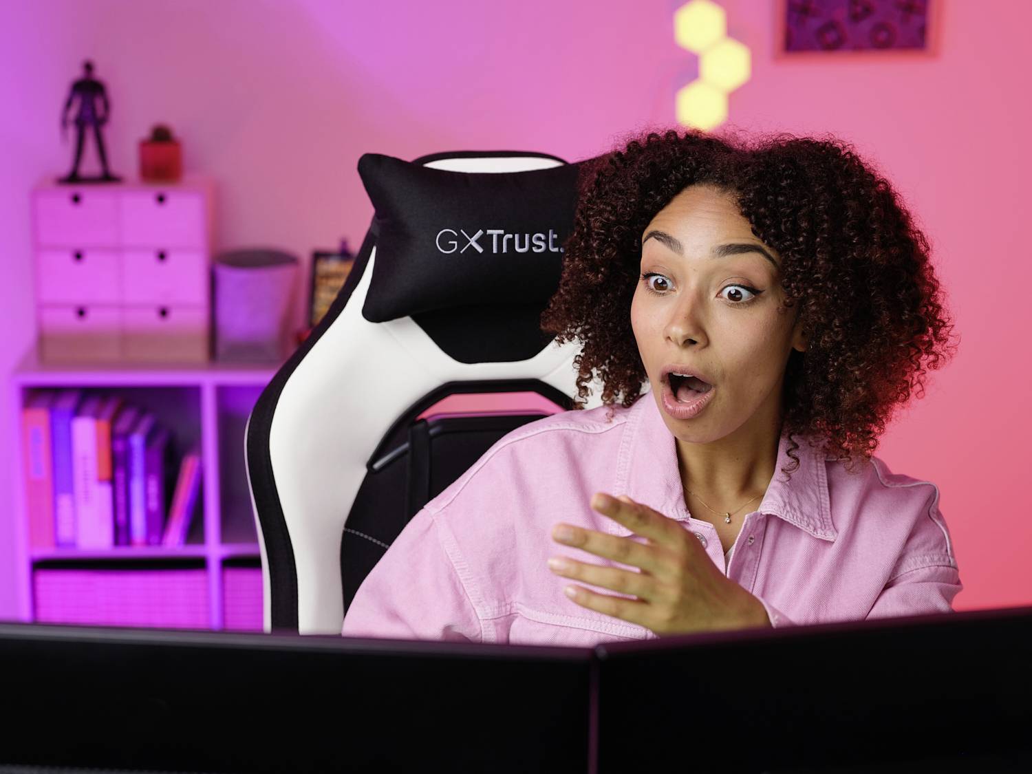 Eine Frau sitzt über überrascht vor einem Computerbildschirm in einem bunt beleuchteten Raum. Im Hintergrund sind Regale mit Büchern.