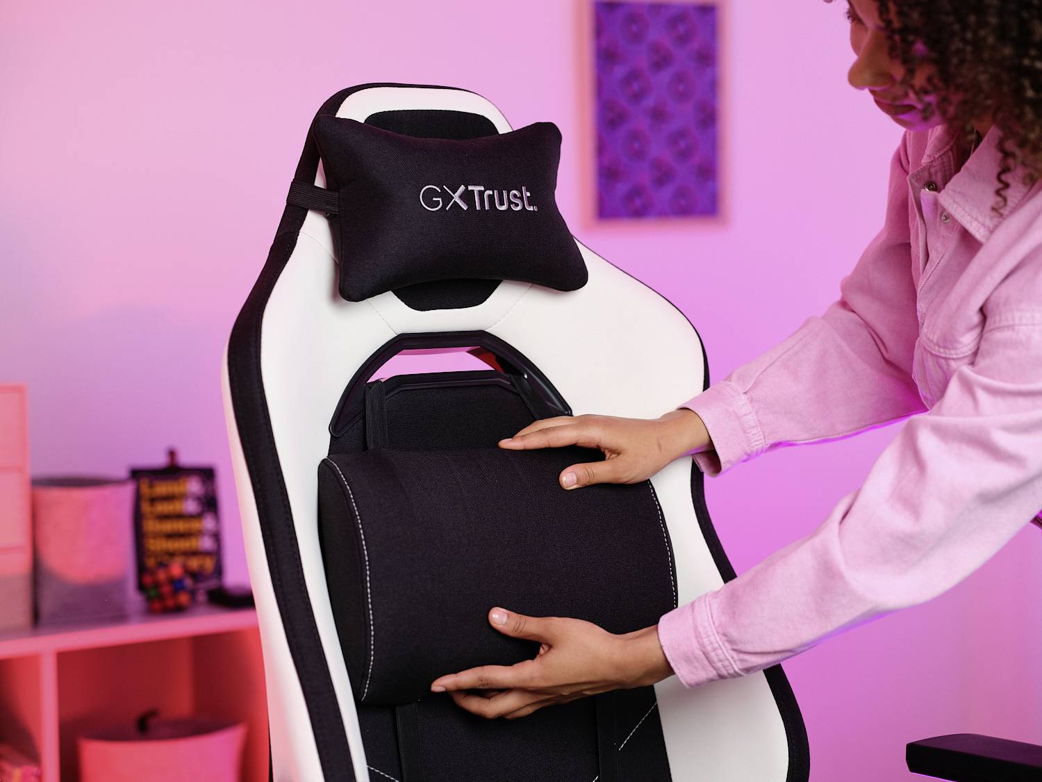 Eine Person justiert ein schwarzes Lendenkissen an einem weißen Gaming-Stuhl mit der Aufschrift 'GX Trust' in einem hell erleuchteten Raum.
