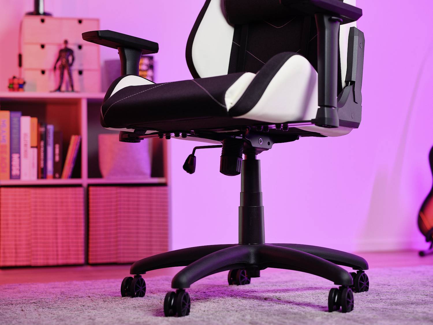 Schwarz-weißer Gaming-Stuhl auf Teppichboden vor Bücherregal und Gitarrenständer, beleuchtet mit rosa Licht.