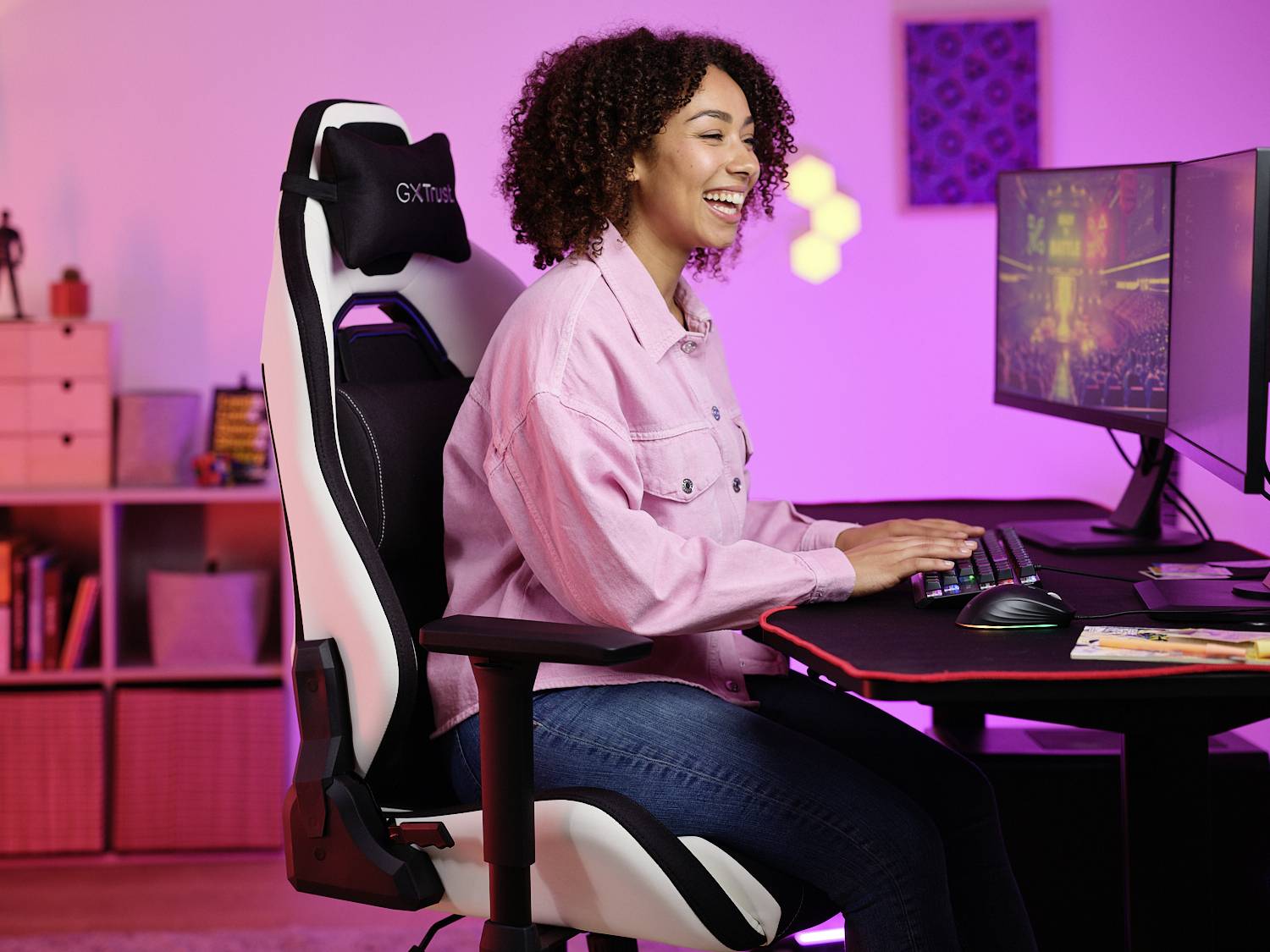 Eine Person sitzt lächelnd an einem Schreibtisch mit zwei Monitoren, spielt oder arbeitet am Computer. Der Raum ist farbenfroh beleuchtet.