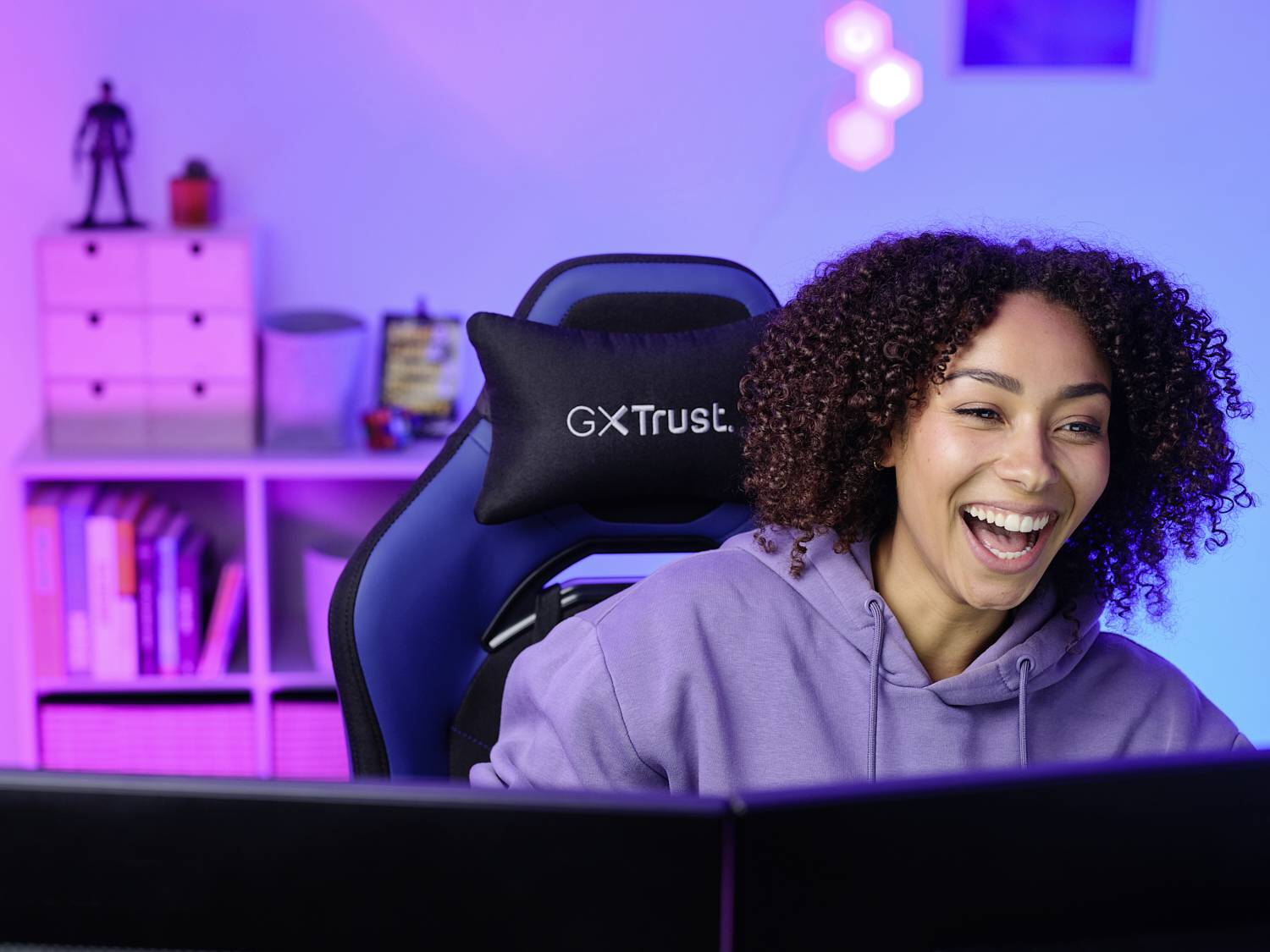 Eine lächelnde Person sitzt vor einem Computer in einem gut beleuchteten Raum mit Regalen und Dekorationen im Hintergrund.