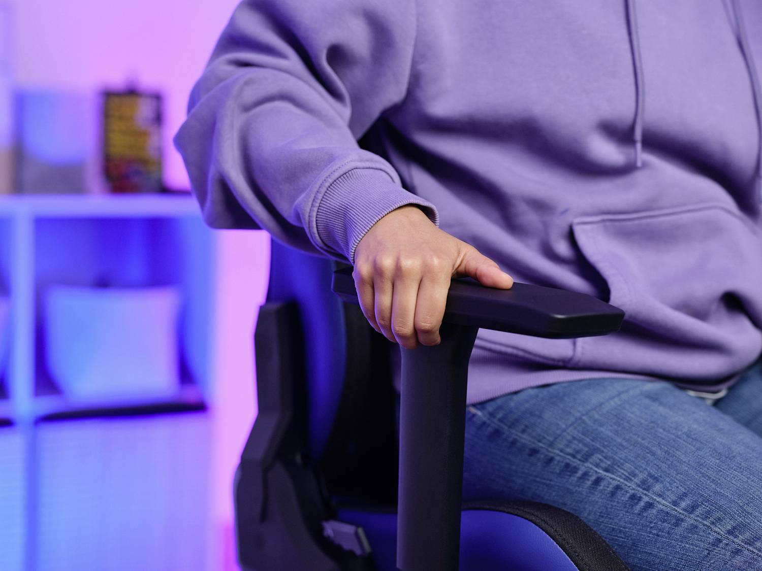 Eine Person in einem lila Pullover sitzt auf einem Stuhl, hält den Armlehne. Im Hintergrund ein leicht unscharfes Regal.