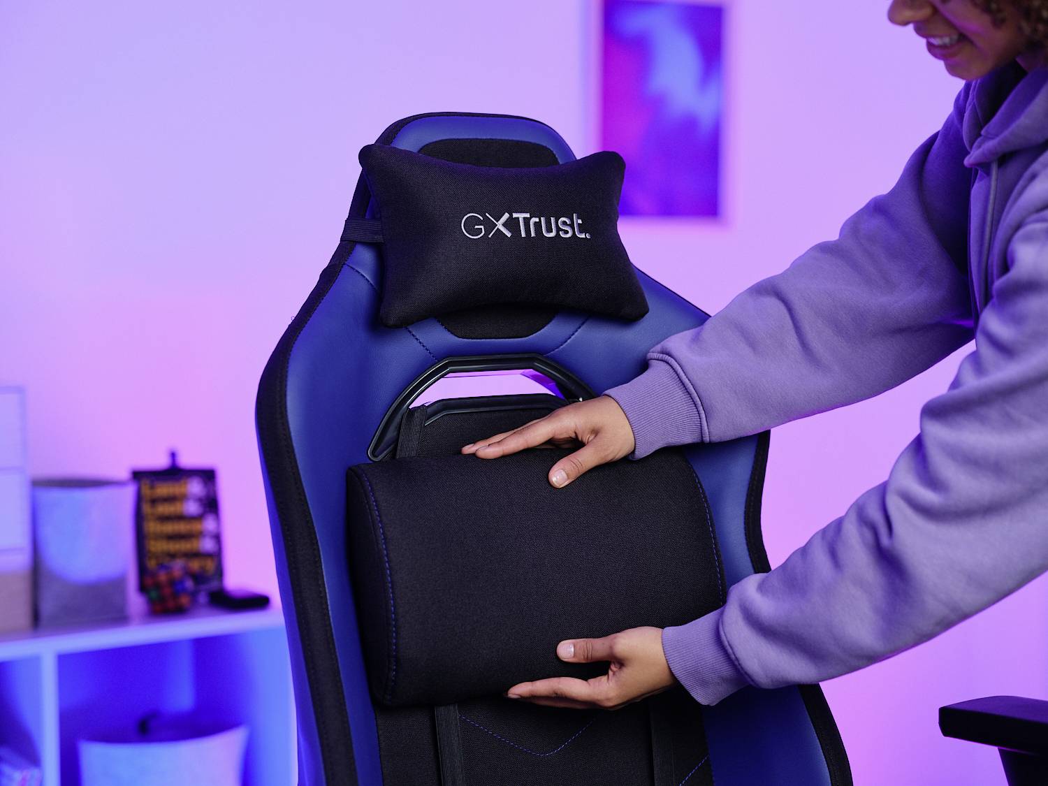 Eine Person justiert das Kissen auf einem blauen GXTrust-Gamingstuhl in einem Raum mit blau-violetter Beleuchtung.