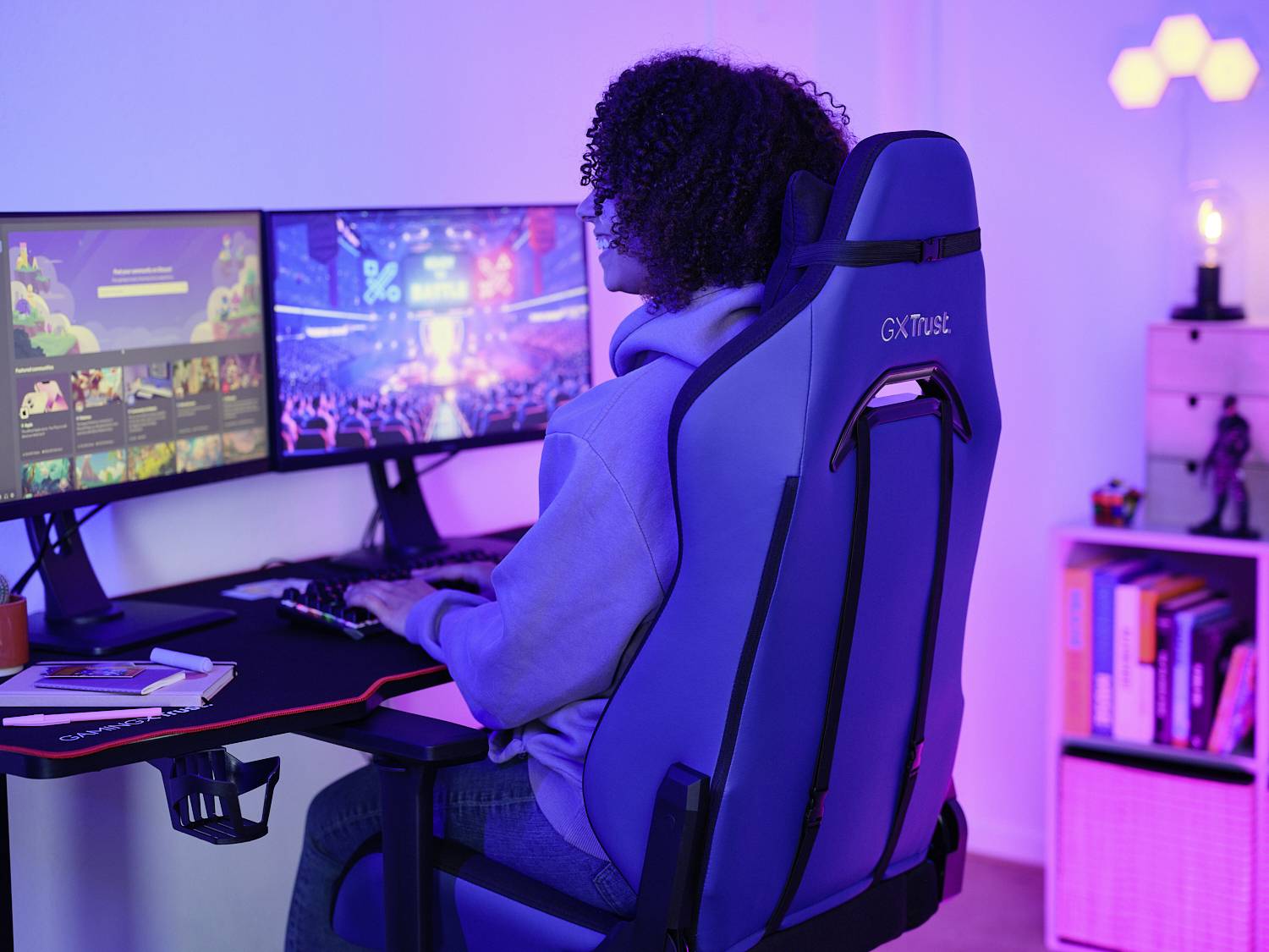 Eine Person sitzt an einem Schreibtisch mit zwei Monitoren, vertieft in ein Computerspiel. Der Raum ist stimmungsvoll blau beleuchtet.