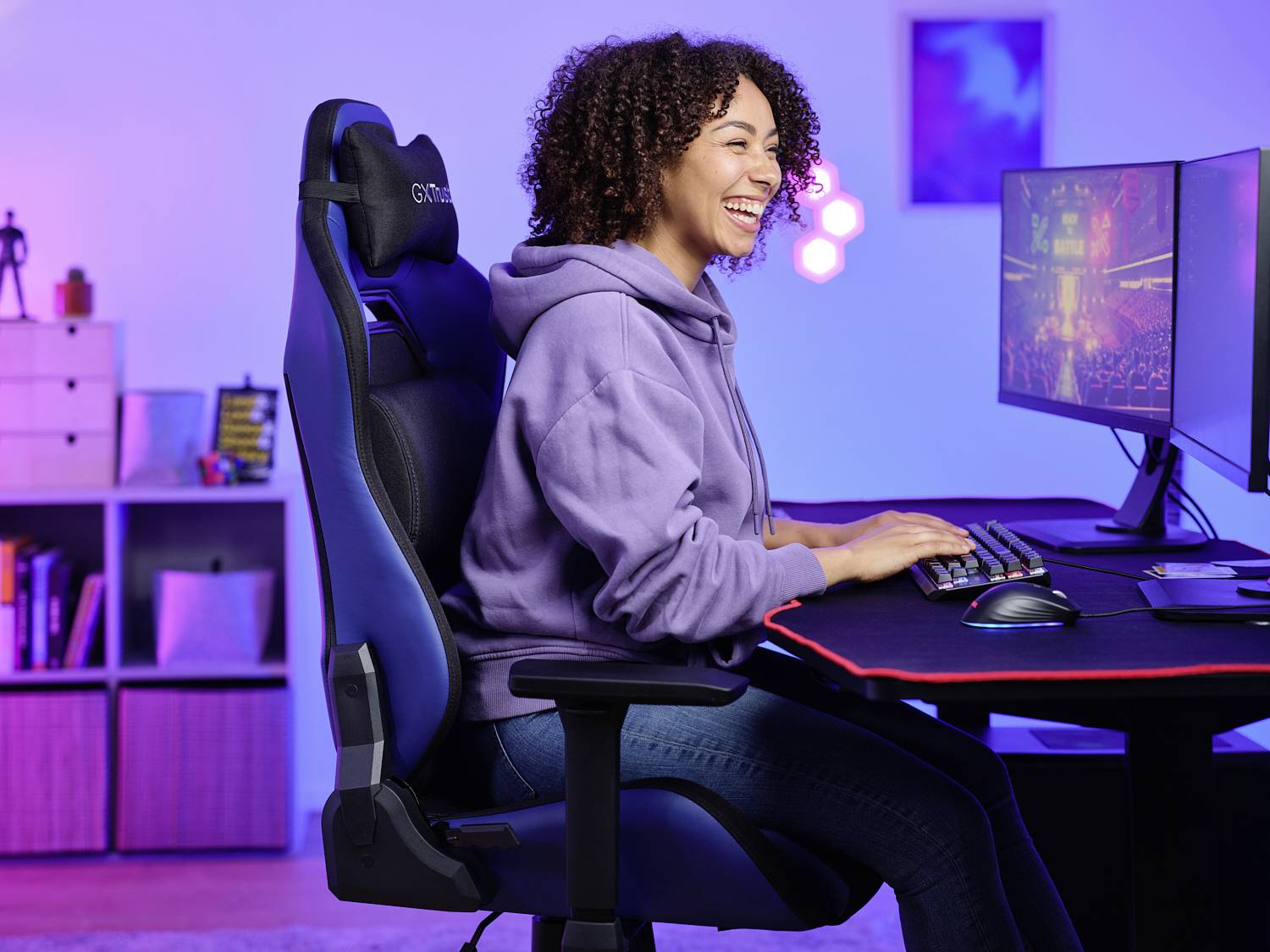 Eine Person sitzt lächelnd in einem Gaming-Stuhl und tippt auf einer Tastatur vor einem Monitor.