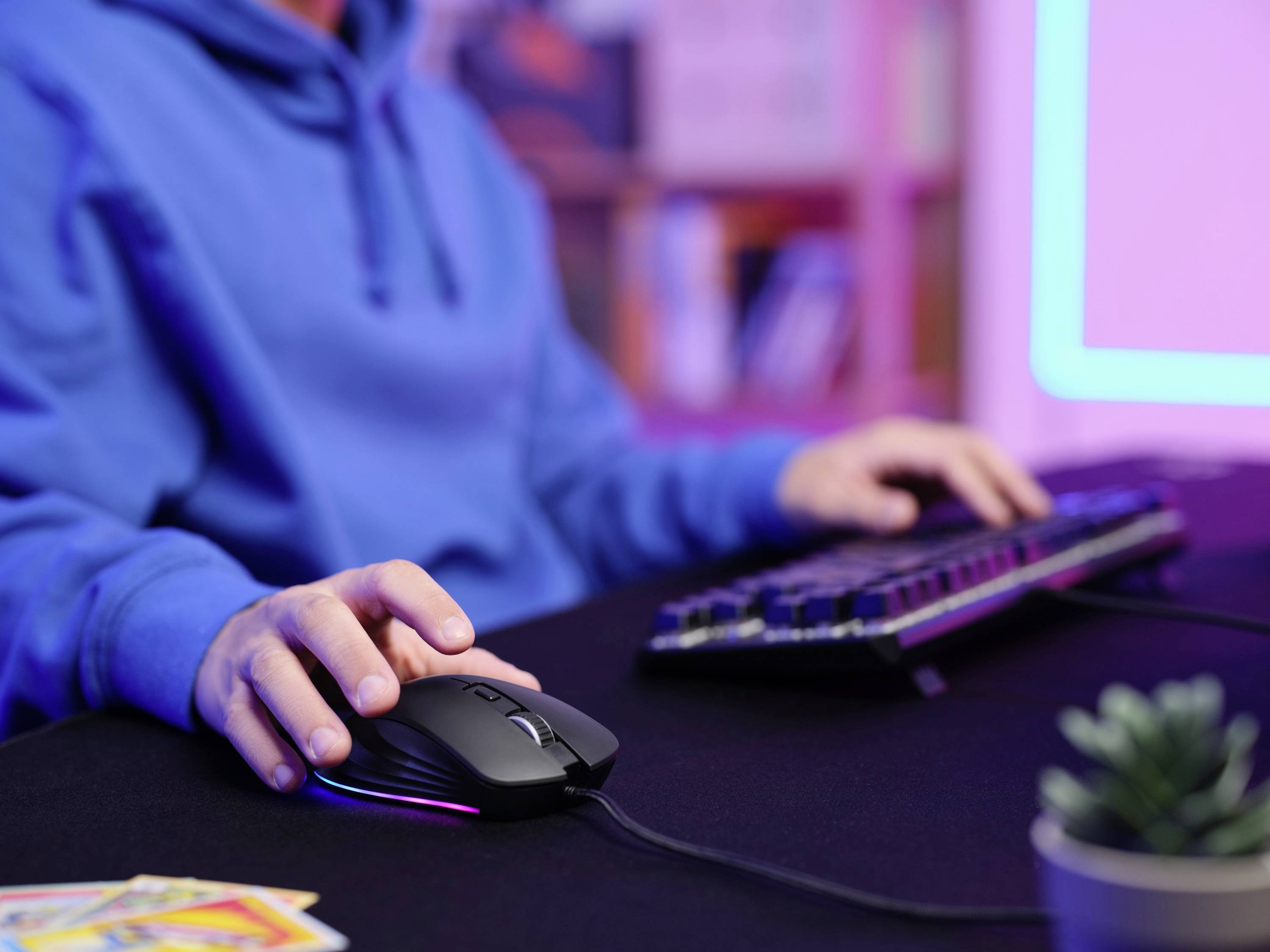 Person in blauem Hoodie benutzt Computermaus und Tastatur an einem Schreibtisch. Im Hintergrund sind Bücherregale zu sehen.