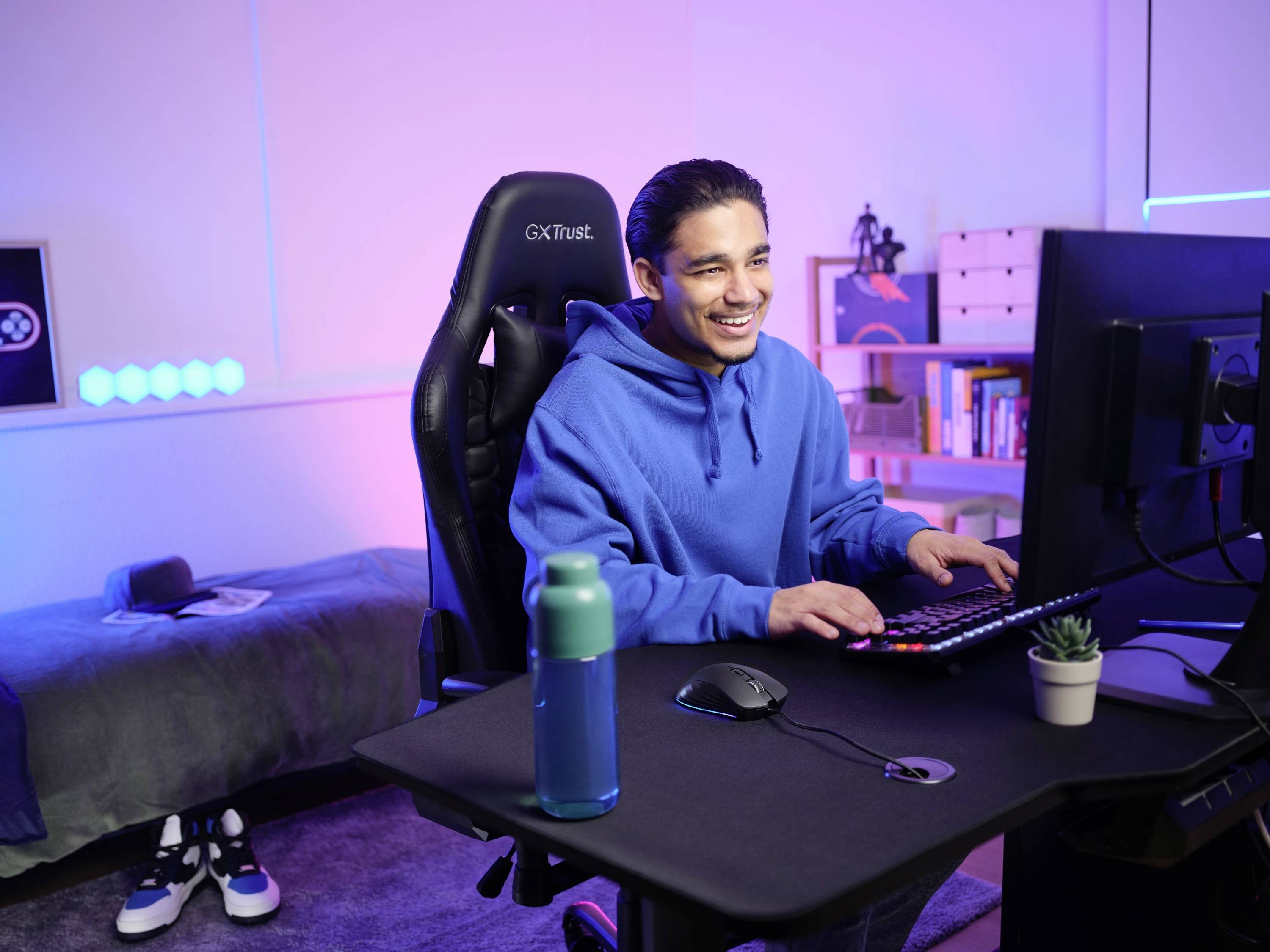 Ein Mann in einem blauen Hoodie sitzt an einem Schreibtisch vor einem Computer und lächelt. Im Hintergrund ist ein Bett und Bücherregal.