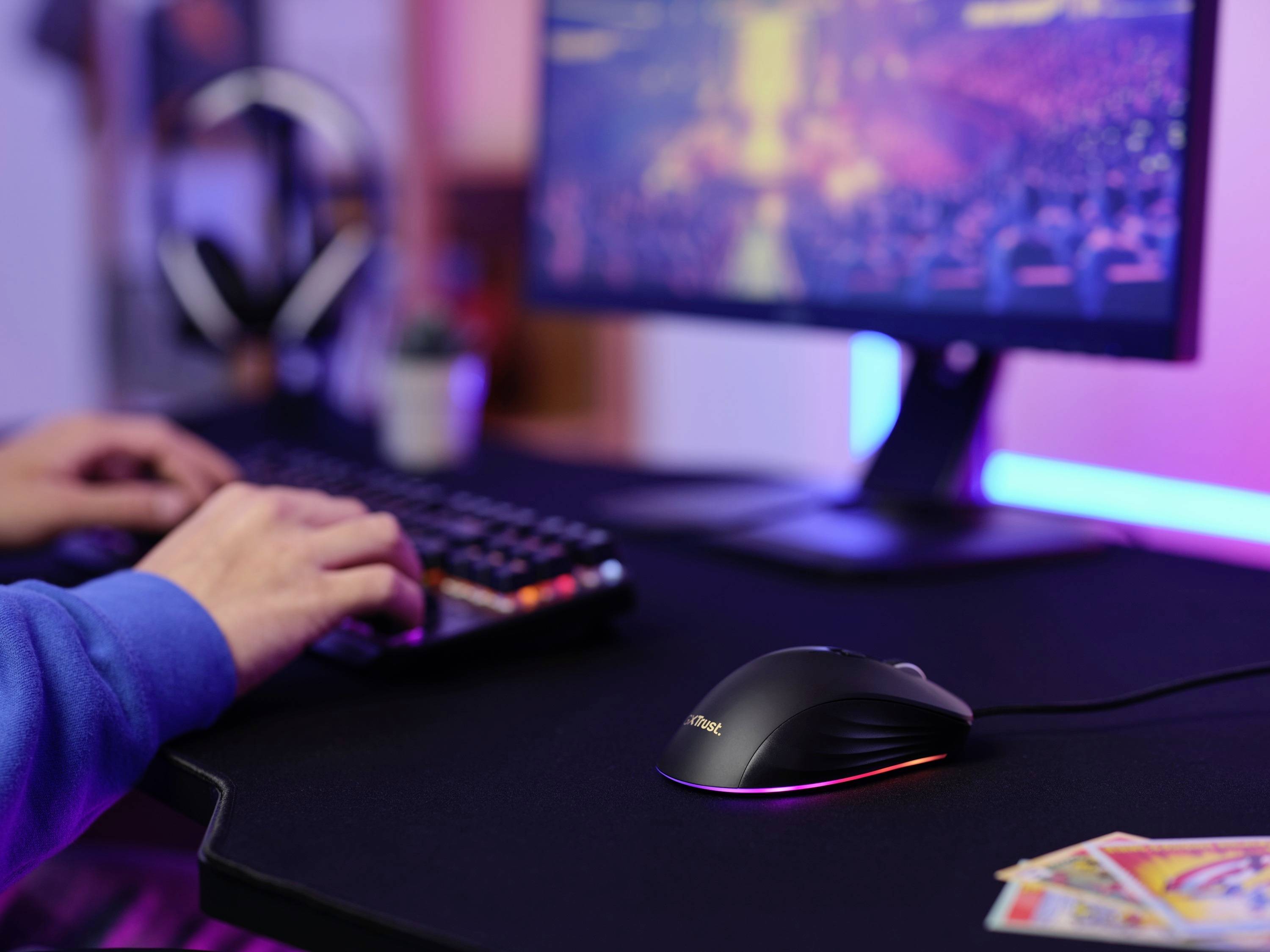 Eine Person tippt auf einer Tastatur vor einem Computerbildschirm, der ein unscharfes Bild anzeigt. Eine Maus und Spielkarten liegen auf dem Tisch.