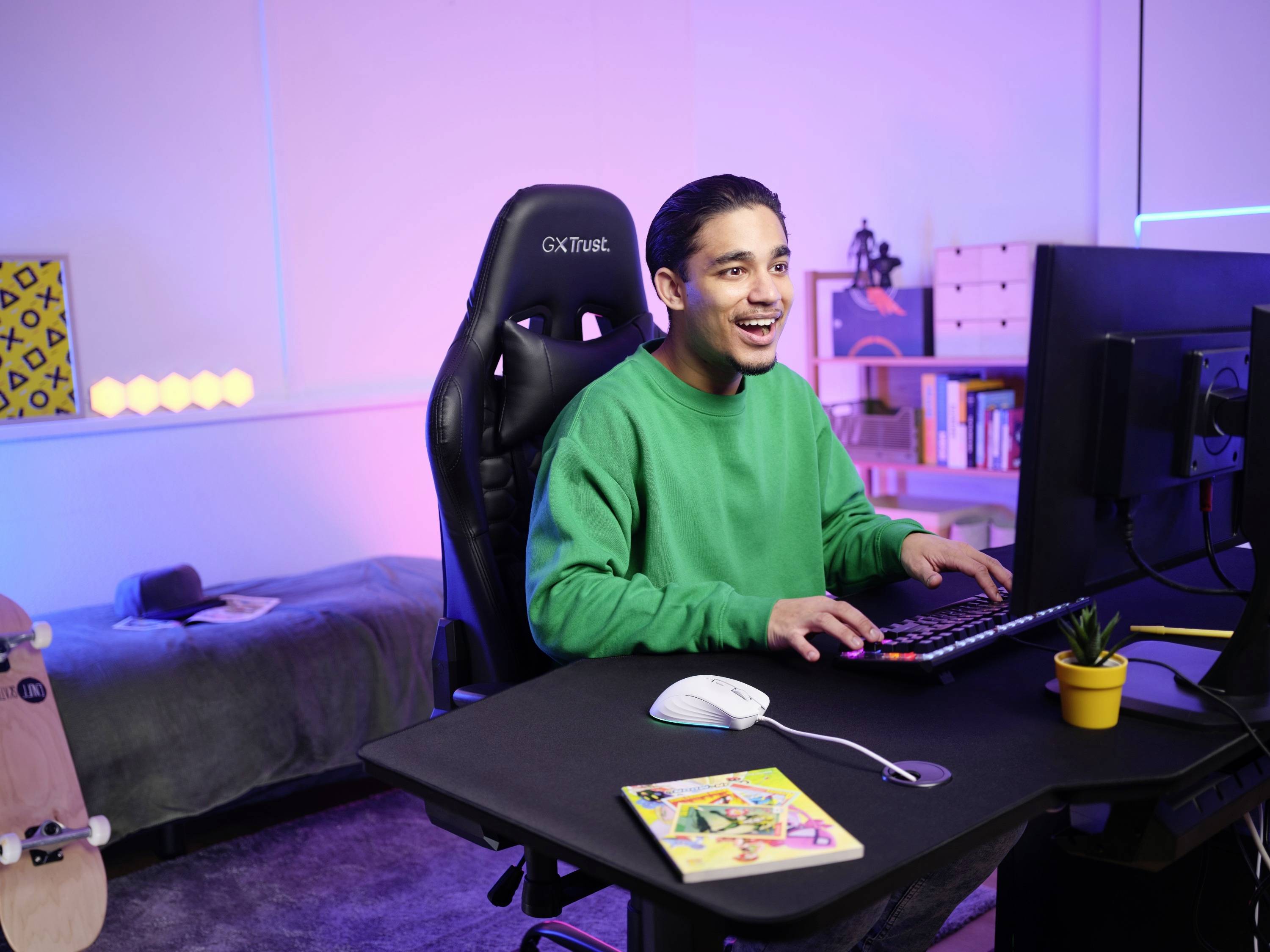 Eine Person sitzt freudig an einem Computer in einem gemütlich eingerichteten Raum mit einem Bett und Regal im Hintergrund.