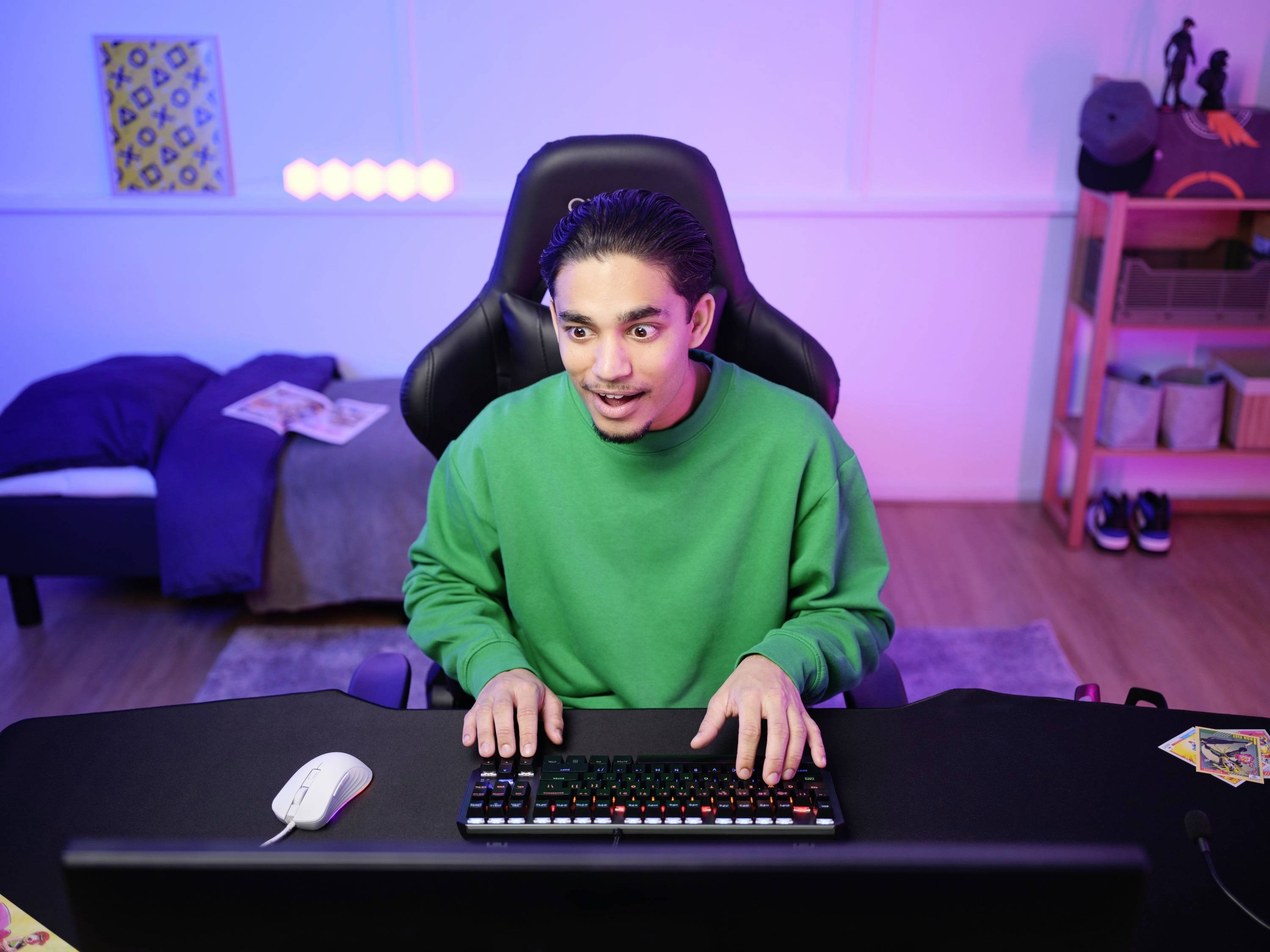 Eine Person in einem grünen Pullover sitzt vor einem Computer und tippt, umgeben von einem beleuchteten Zimmer, das ein Schlafzimmer zu sein scheint.