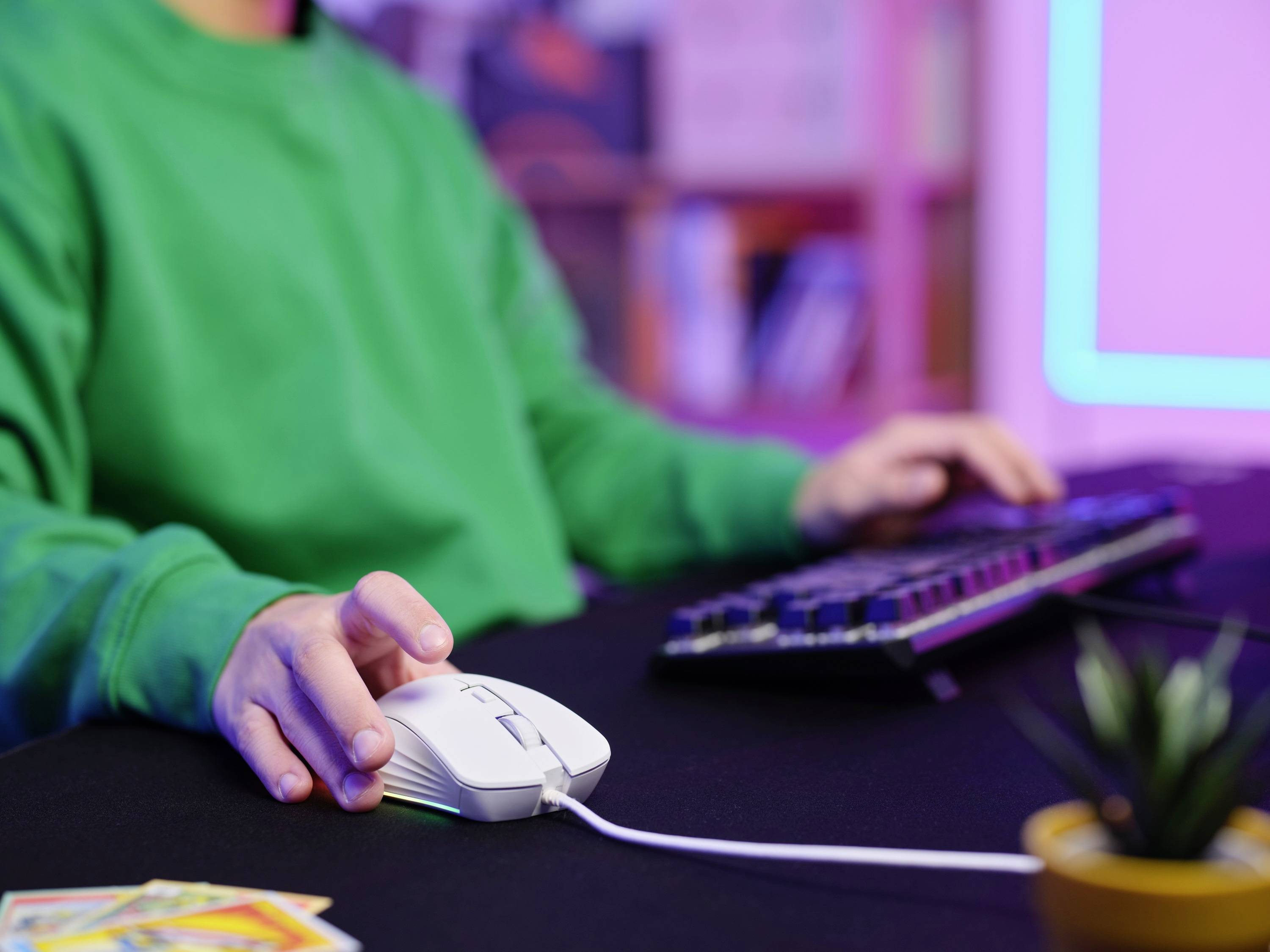 Eine Person in einem grünen Pullover benutzt eine Computermaus und tippt auf einer Tastatur in einem farbenfrohen Raum.